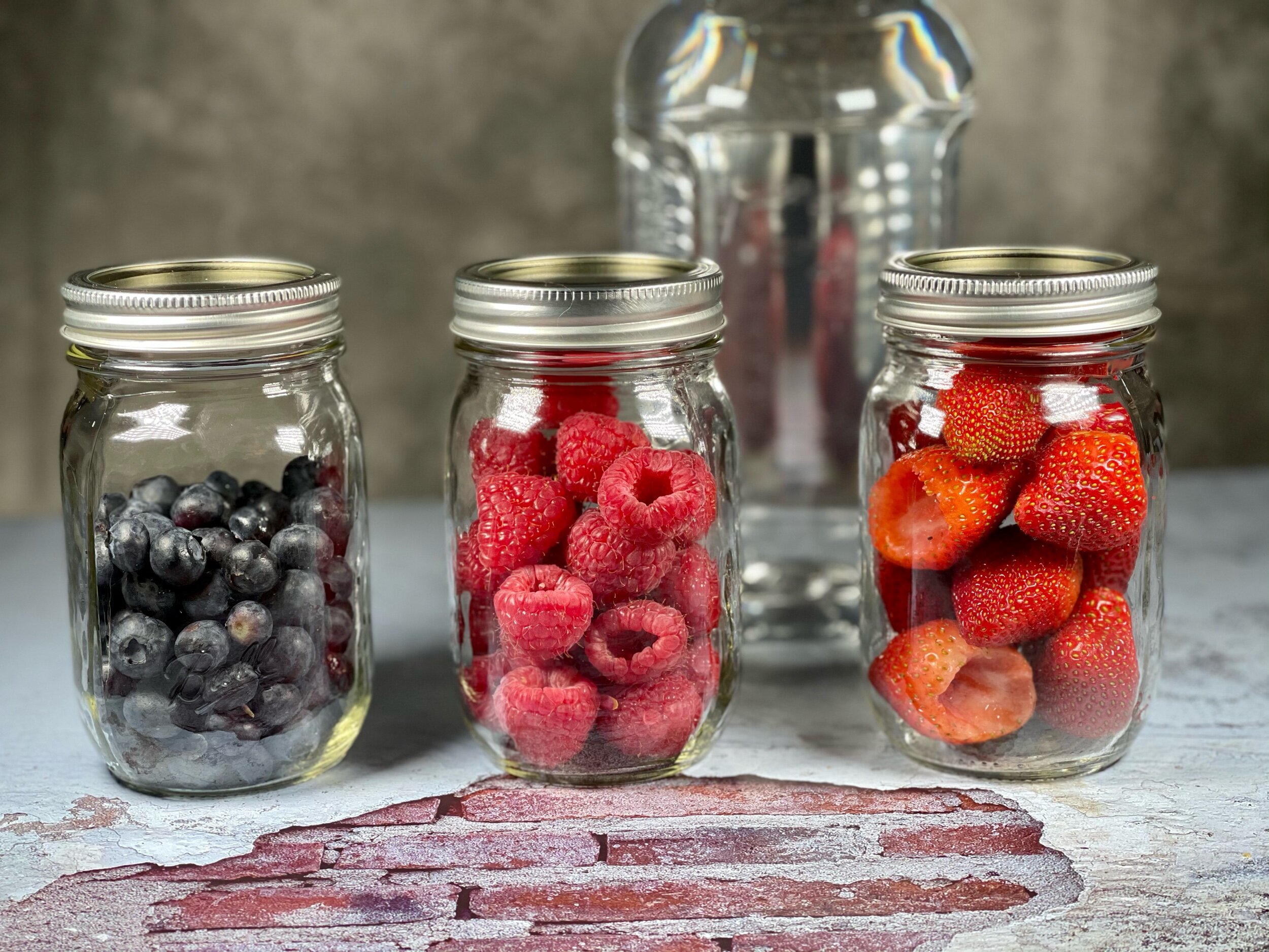  Left to right close up of a jar of blueberries , raspberries , and strawberries with a bottle of vodka placed behind the jars. 