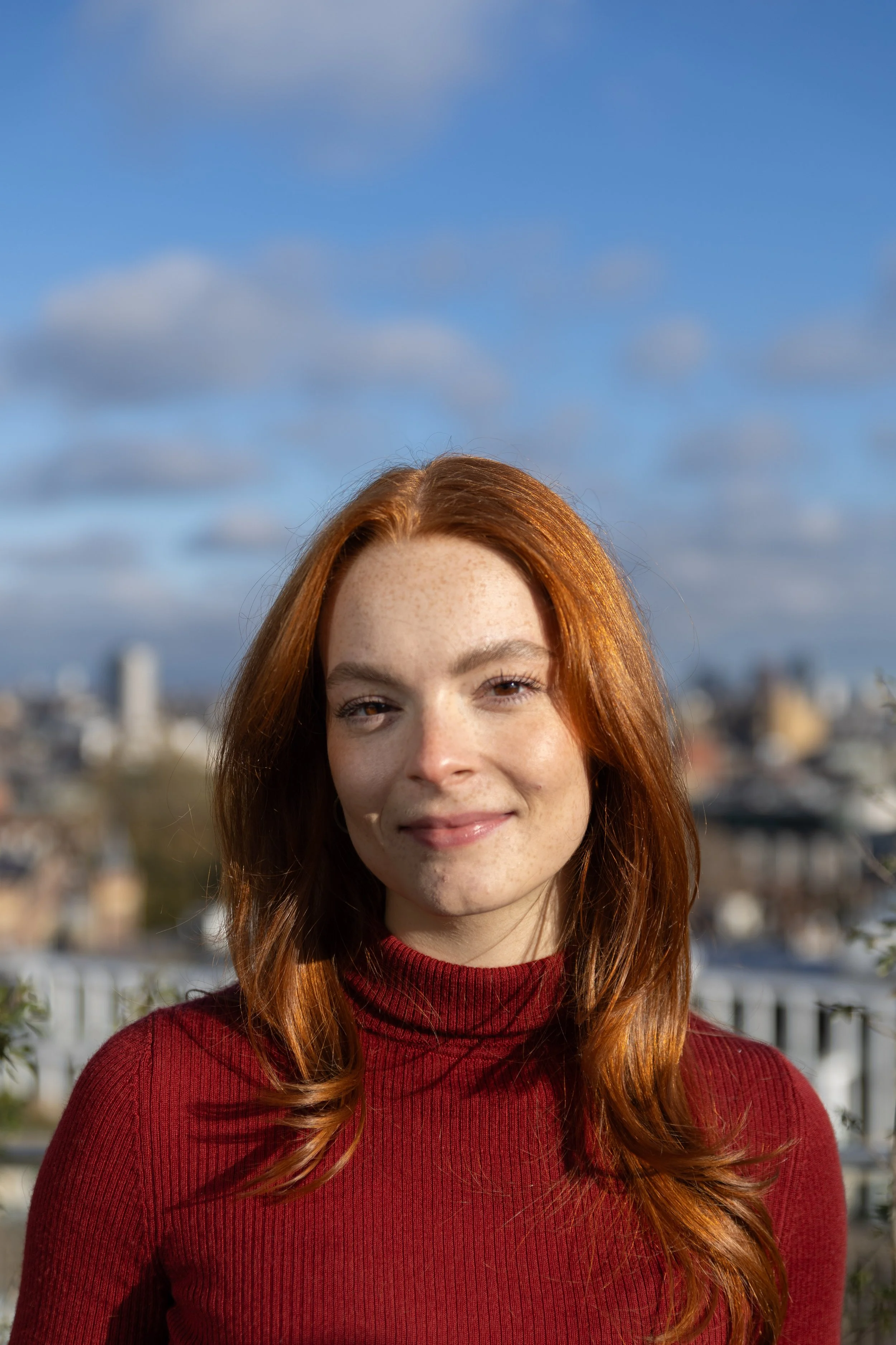 A young woman with long brown hair and blue eyes, wearing a dark coat and green turtleneck, standing outdoors on a cloudy day with city buildings in the background.