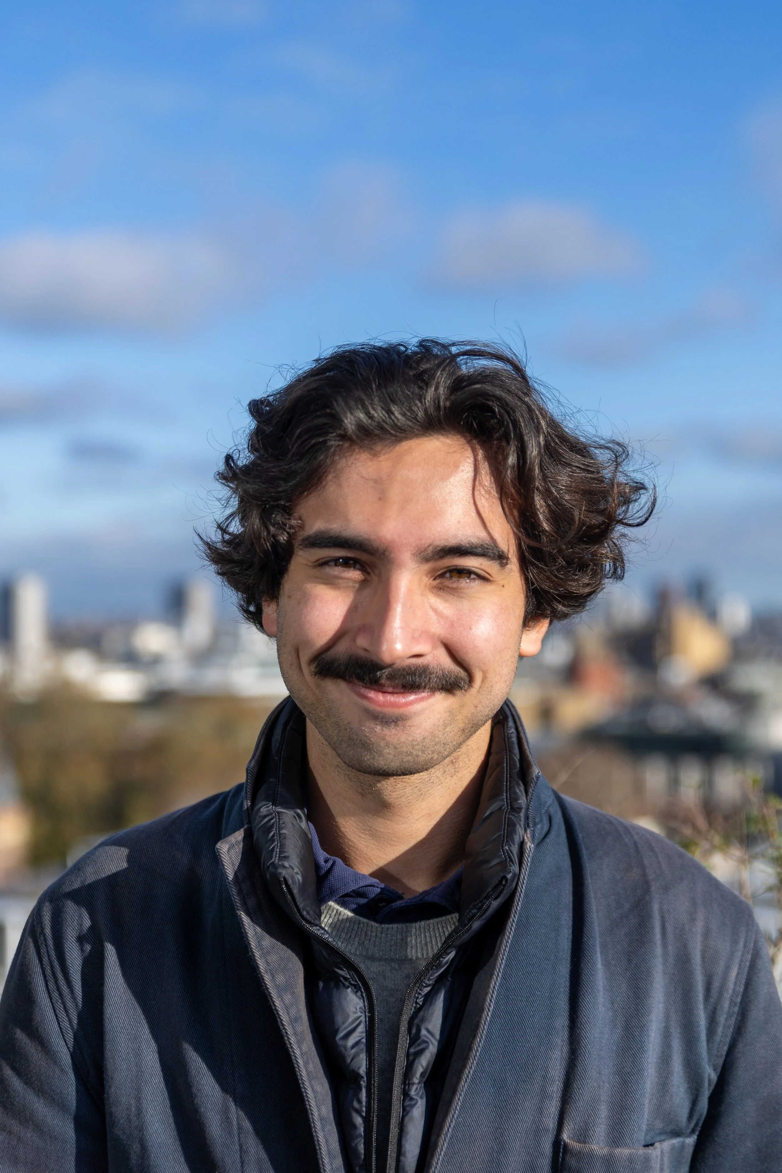 A young man smiling outdoors on a city rooftop with skyscrapers in the background.