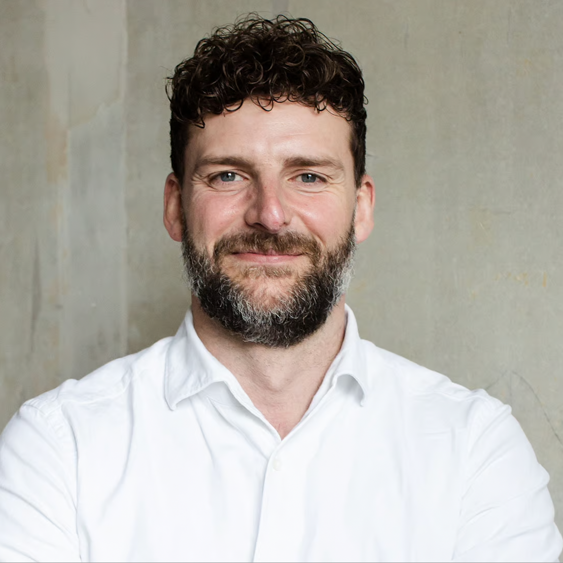 Portrait of a man with curly brown hair and a beard, smiling, wearing a white shirt, standing against a neutral background.