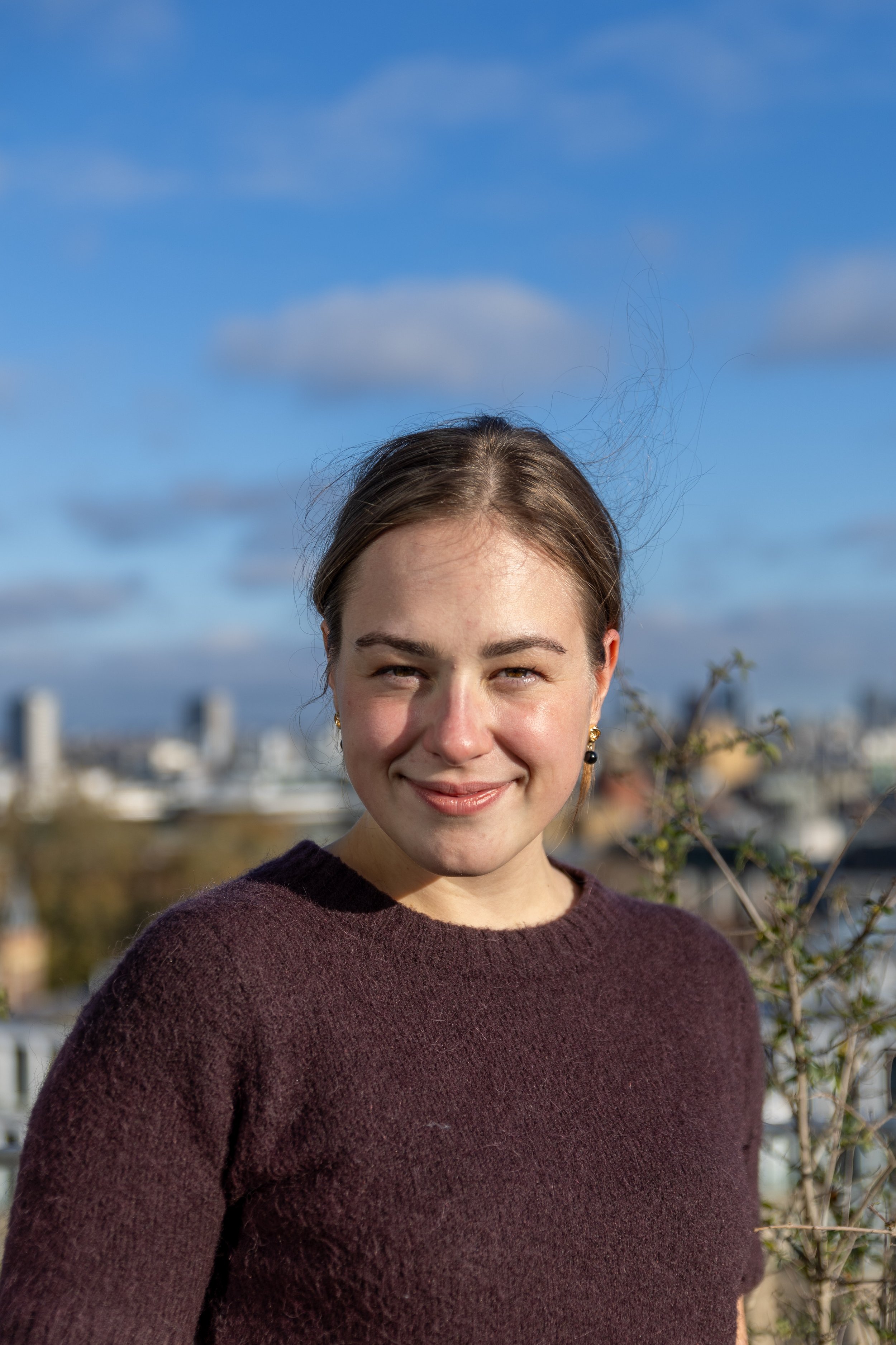 A young man with brown hair and fair skin, wearing a brown and white striped sweater, standing on a rooftop with a city skyline in the background, on an overcast day.