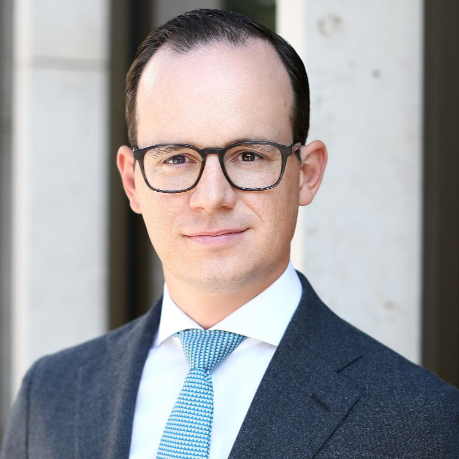 A man in a business suit with glasses, white shirt, and a blue patterned tie, standing outdoors in front of a building.
