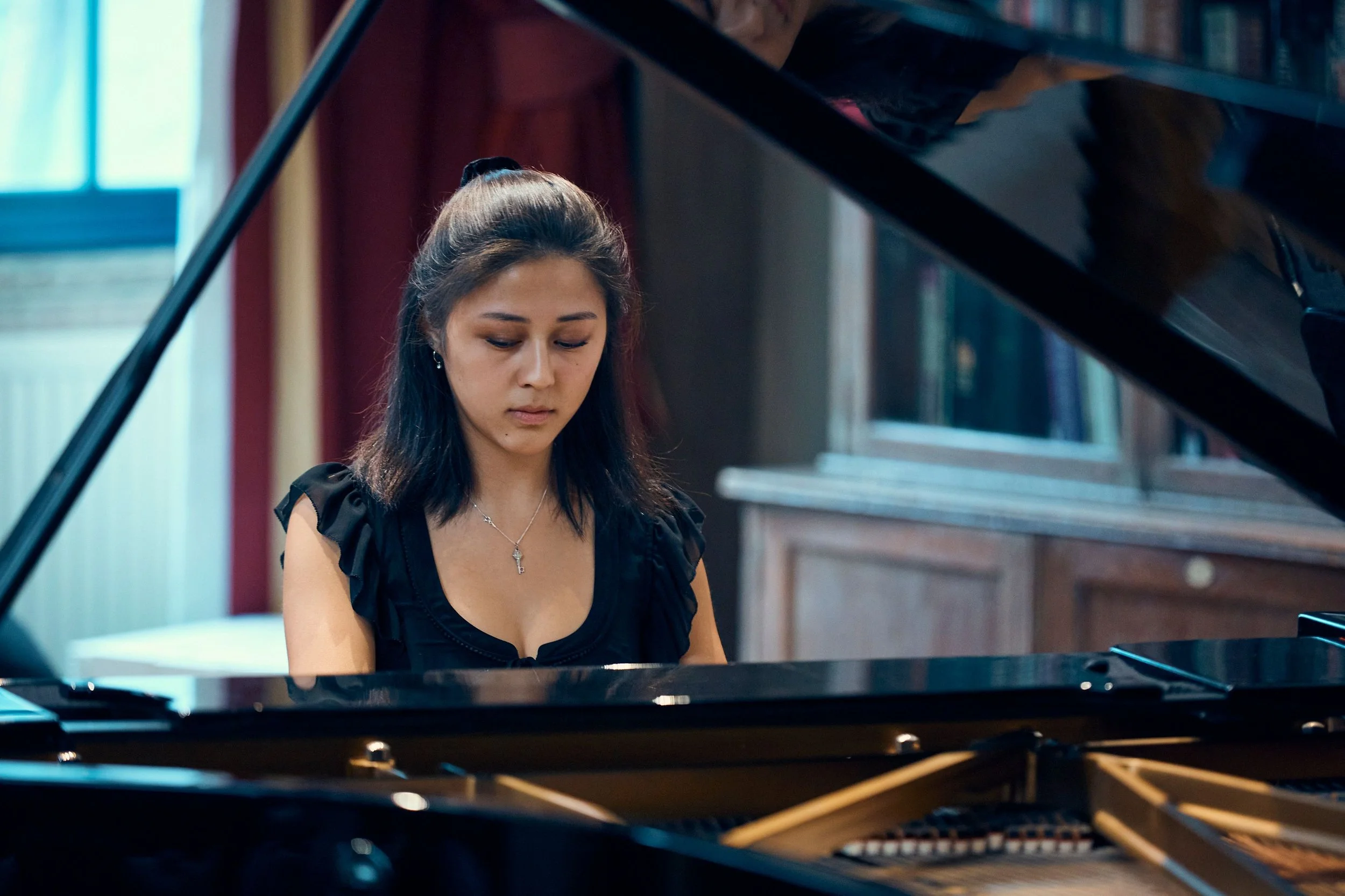 Young woman playing a grand piano in a room with wooden furniture and a window.
