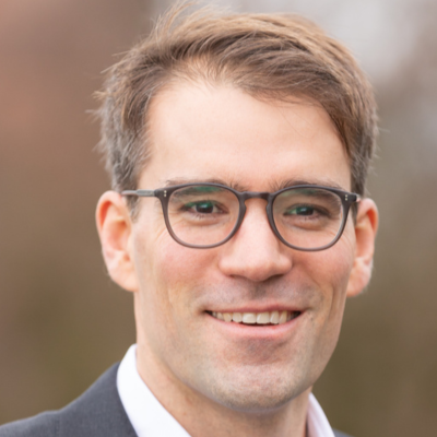 Close-up of a man in glasses smiling outdoors with a blurred natural background.