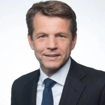 A professional headshot of a man with brown hair, wearing a dark suit, white shirt, and blue tie, smiling against a plain light background.