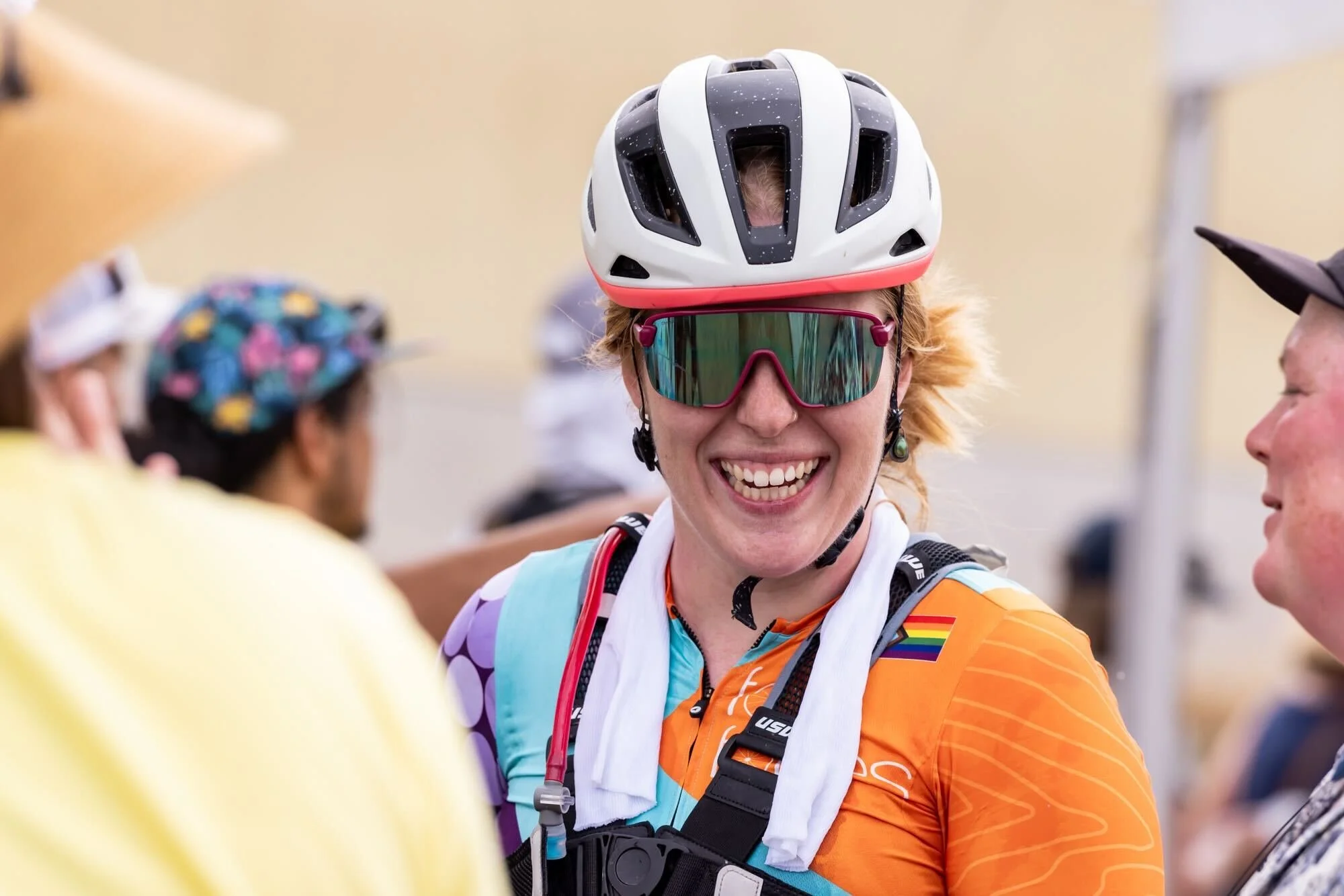 A smiling woman wearing a white bike helmet, sports sunglasses, and a colorful Follies cycling jersey standing among a group of people at the end of a race.