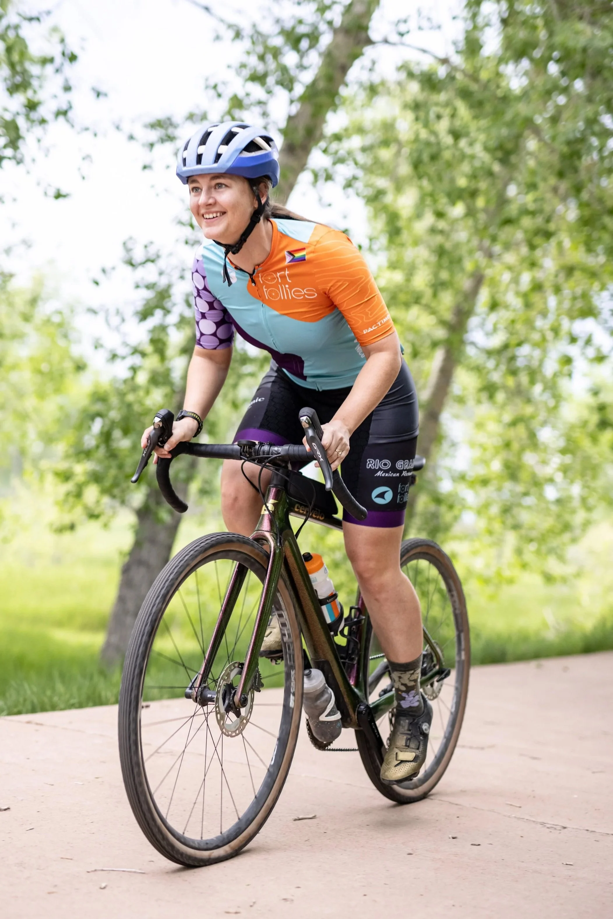 A woman riding a black road bike on a paved path in a park, smiling and wearing a colorful cycling jersey, black shorts, a bike helmet, and cycling shoes, with trees and greenery in the background.
