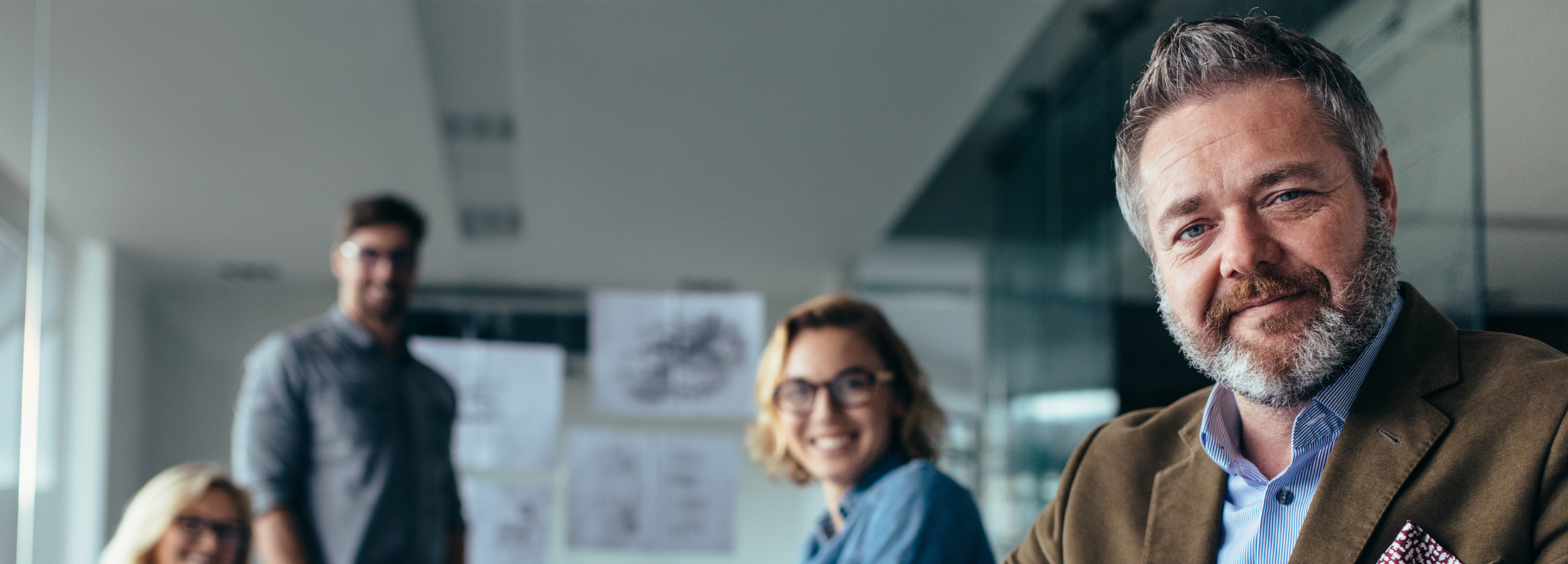 A group of four professionals smiling in a modern office, with a focus on a smiling middle-aged man with gray hair and a beard in the foreground.