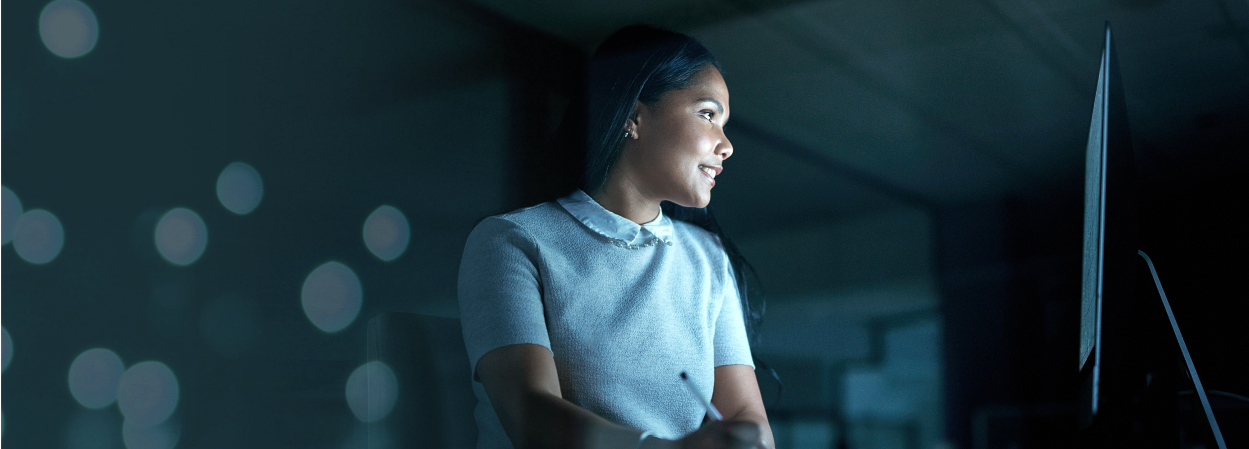Woman working at a computer in a dimly lit office with bokeh lights in the background.
