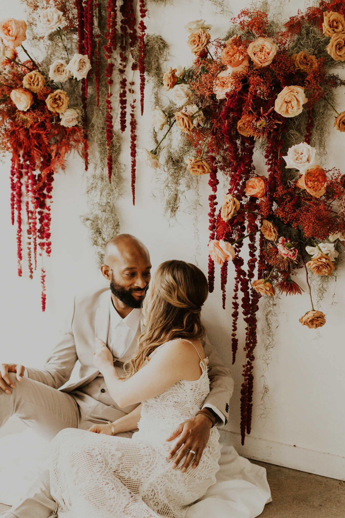  This recently married couple poses in love for a boho inspired style wedding photoshoot in Denver, Colorado captured by Dawn Louise Photography. Bride and groom floral backdrop boho inspired wedding greenery flowers white lace wedding dress tan groo