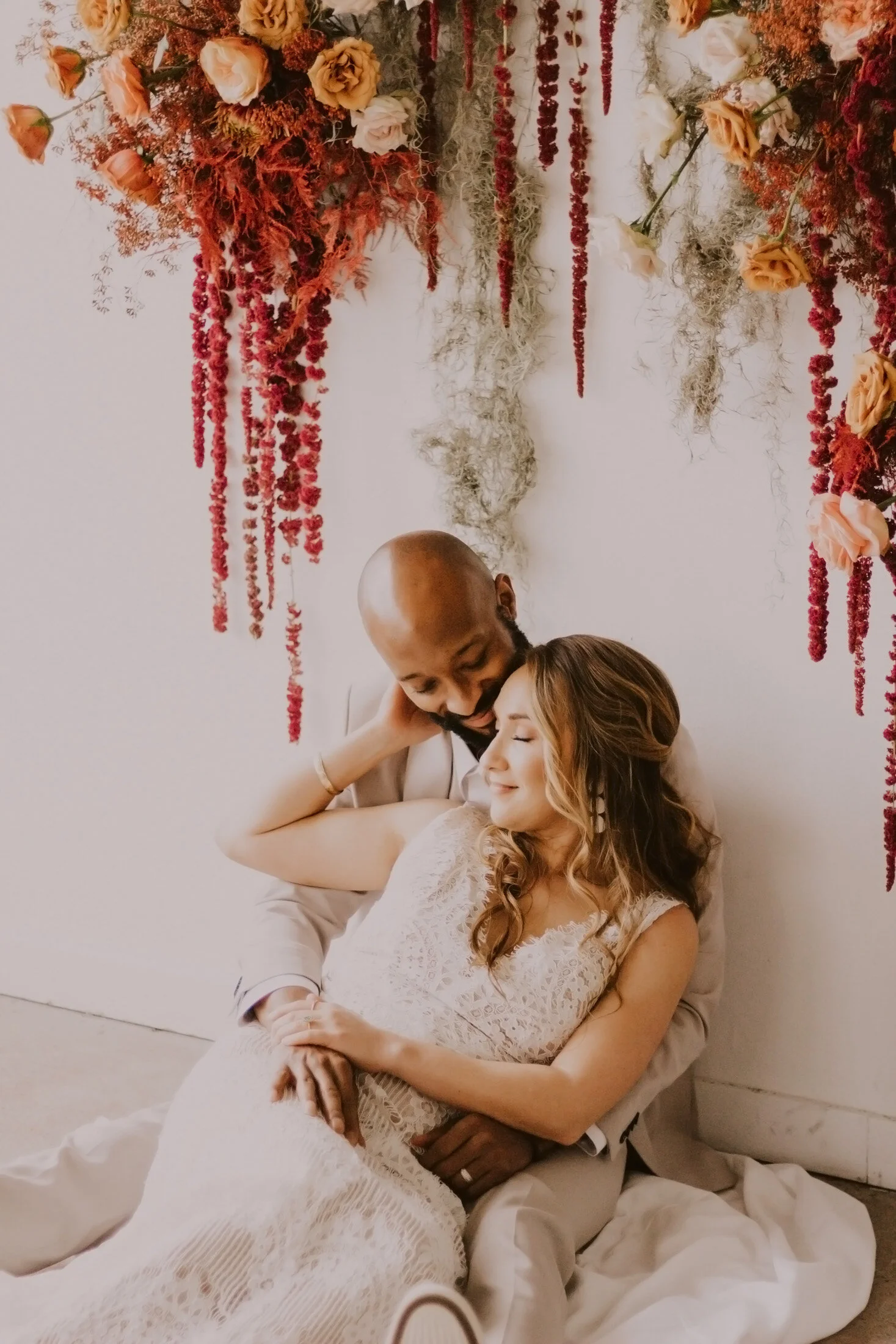  Bride and groom sit and pose together in a stunning boho inspired styled wedding photoshoot with Dawn Louise Photography in Denver, Colorado. Dawn Louise photos bride and groom pose inspo boho wedding decor red pink floral backdrop simple elegant we