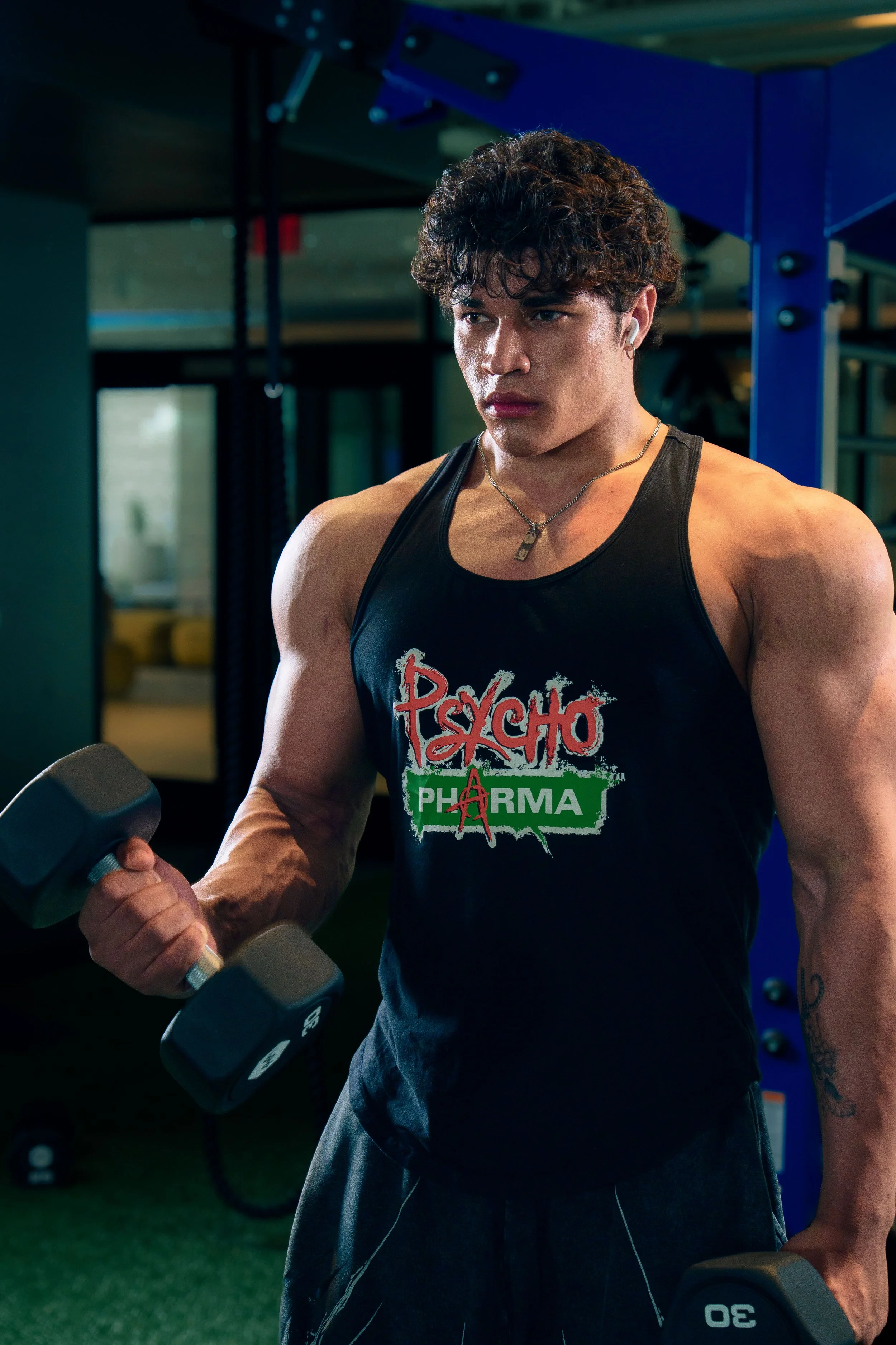 Young man with curly hair wearing a black tank top and exercising in a gym, holding a dumbbell.