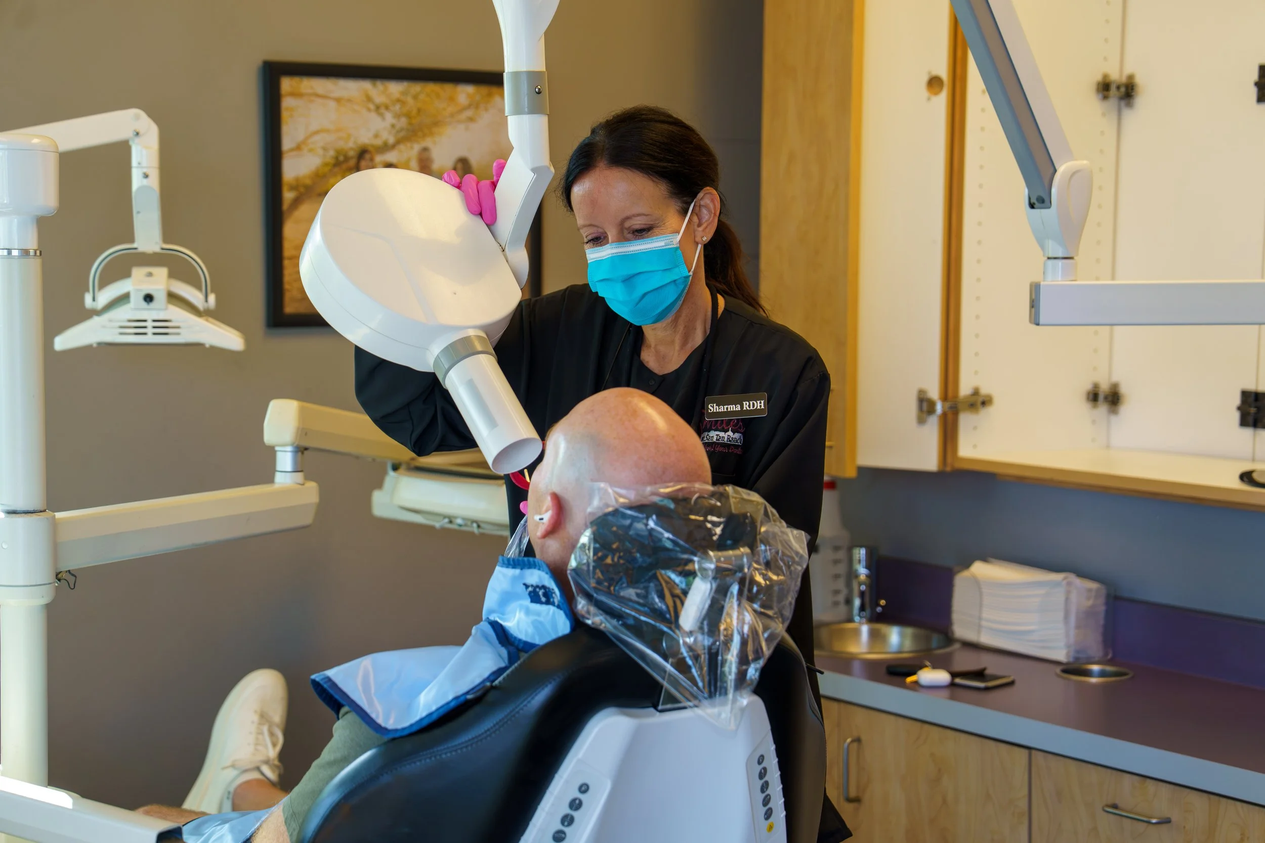 A healthcare professional wearing a face mask and gloves examining a patient's teeth or mouth using dental equipment in a clinic setting.