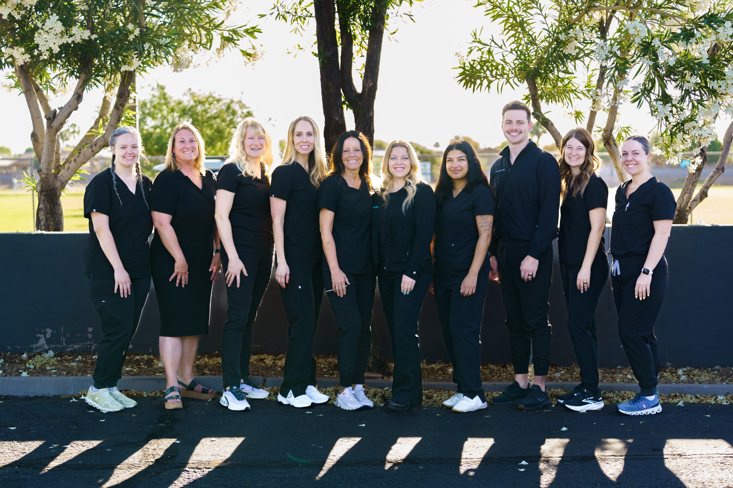 Group of nine people standing outdoors in front of trees with white flowers, smiling for the camera, wearing black shirts and casual pants.