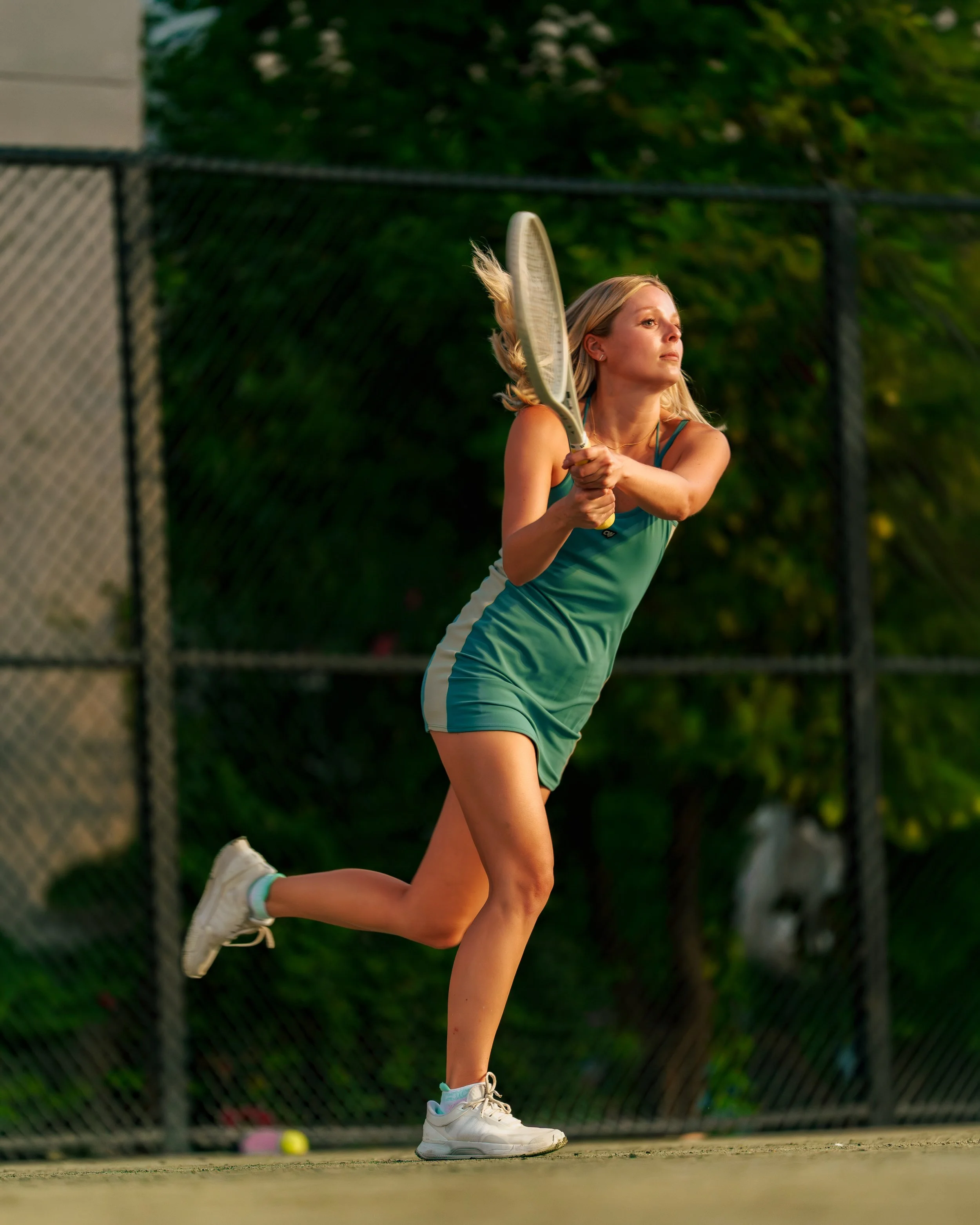 A woman with blonde hair playing tennis on a court, holding a tennis racket, with a tennis ball on the ground nearby, wearing a teal sports dress and white sneakers.