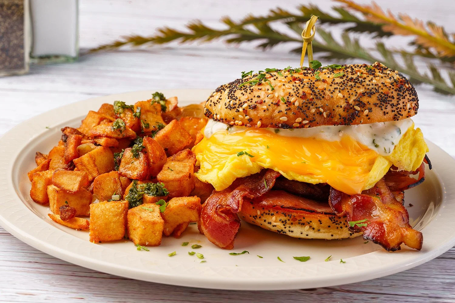 Plate of breakfast with a bacon, egg, and cheese sandwich with poppy seed and sesame seed bagel, served with roasted potatoes garnished with herbs.