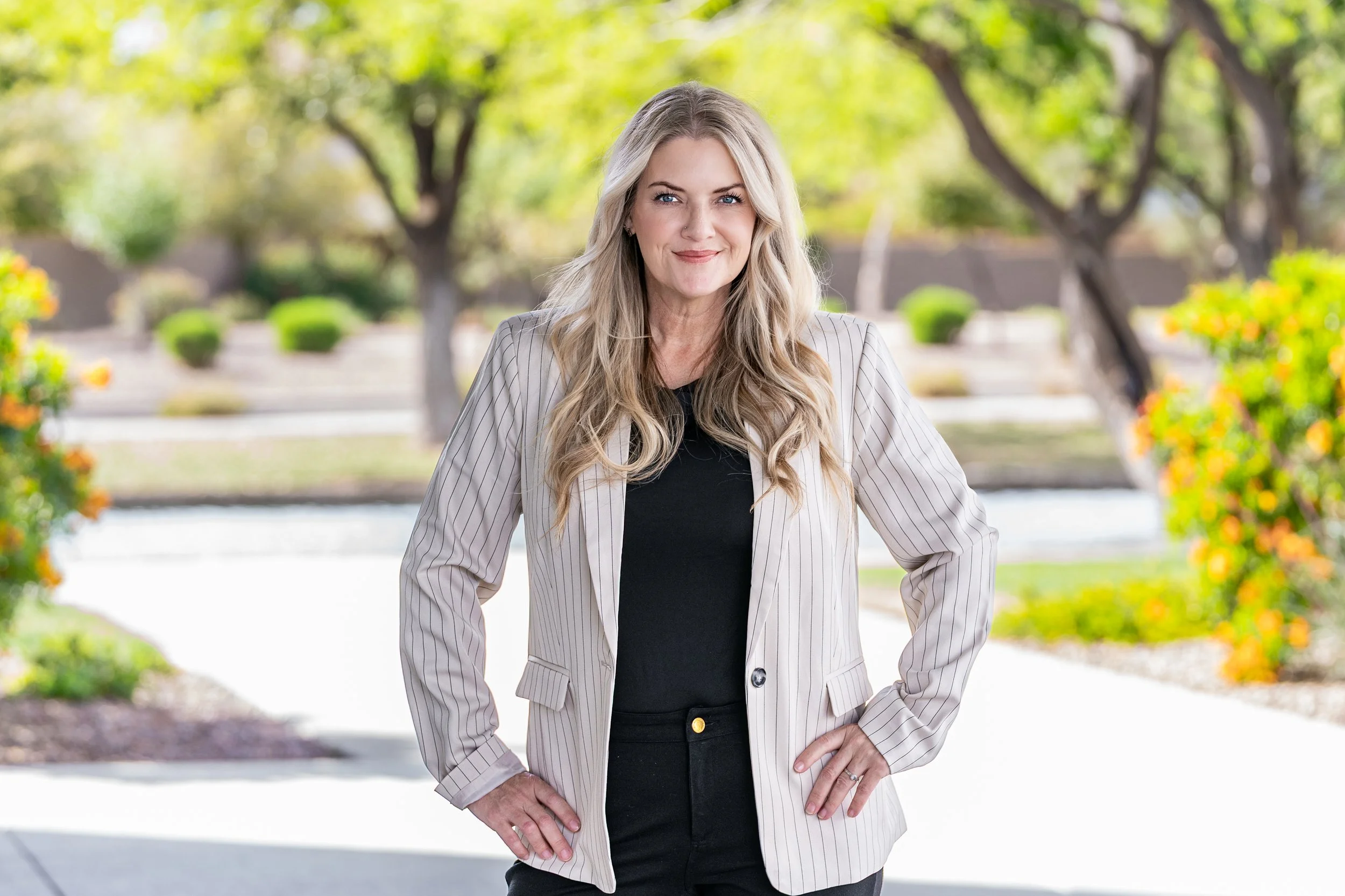 A woman with long blonde hair smiling outdoors in a sunny park or garden, wearing a light striped blazer and black top.
