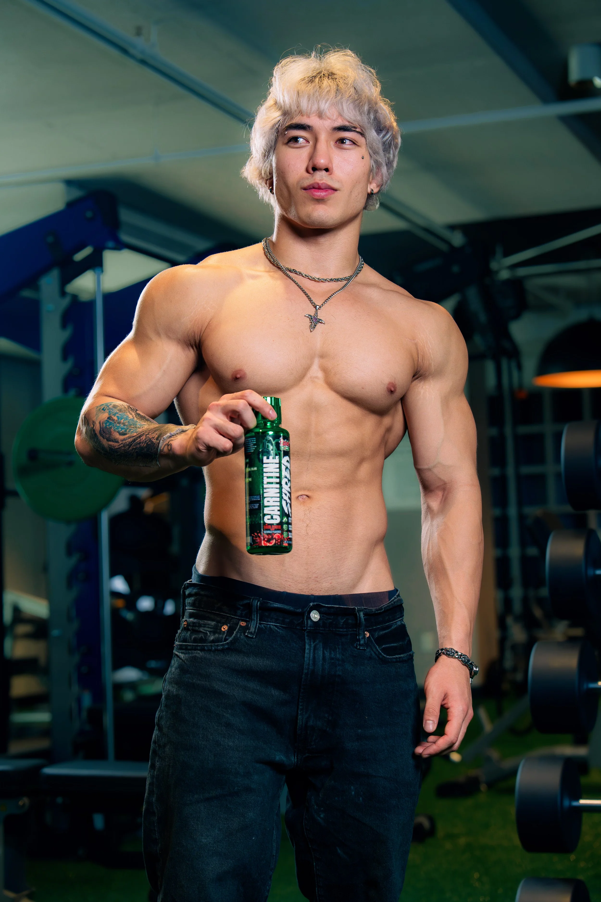 A young shirtless man with platinum blonde hair, tattoos on his left arm, wearing chains, stands in a gym holding a green bottle labeled 'CARNITINE'.