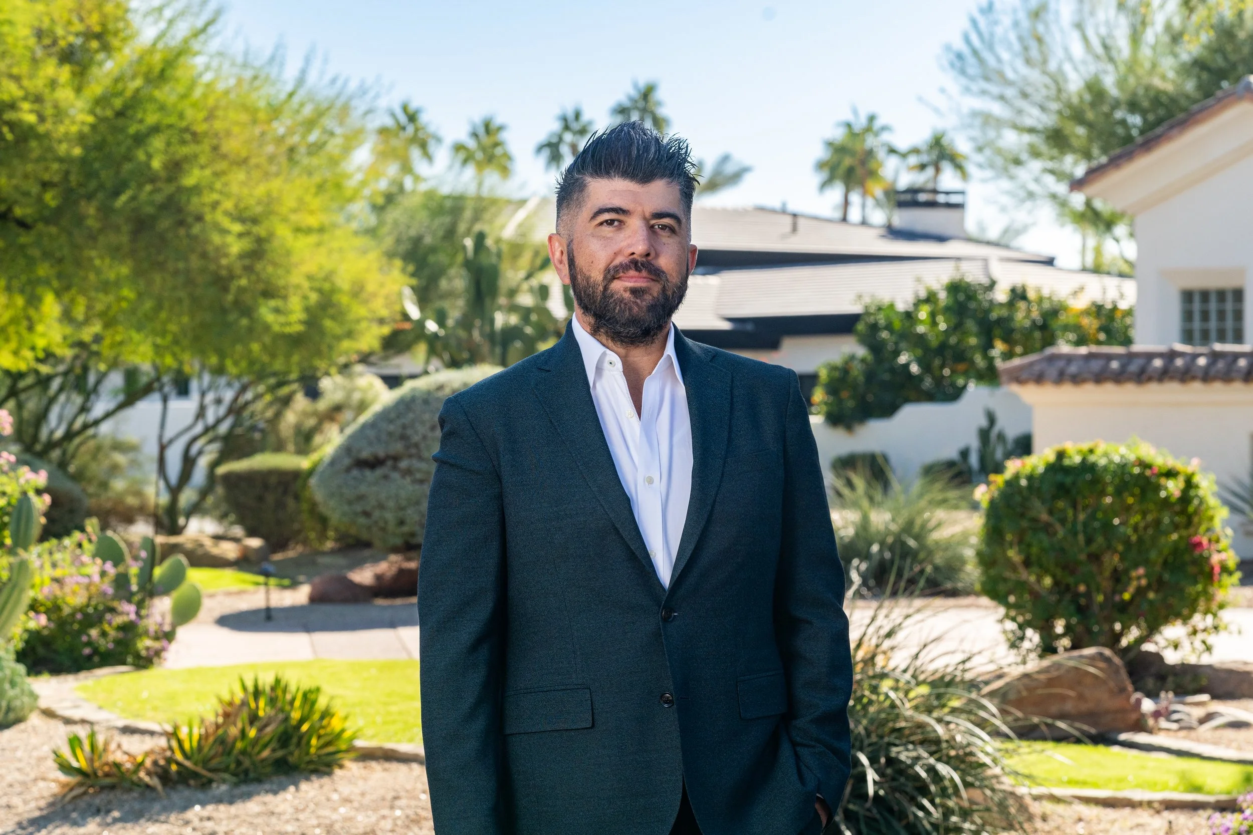 A man in a dark suit and white shirt standing outdoors in front of a well-kept garden with trees, bushes, and a house in the background.