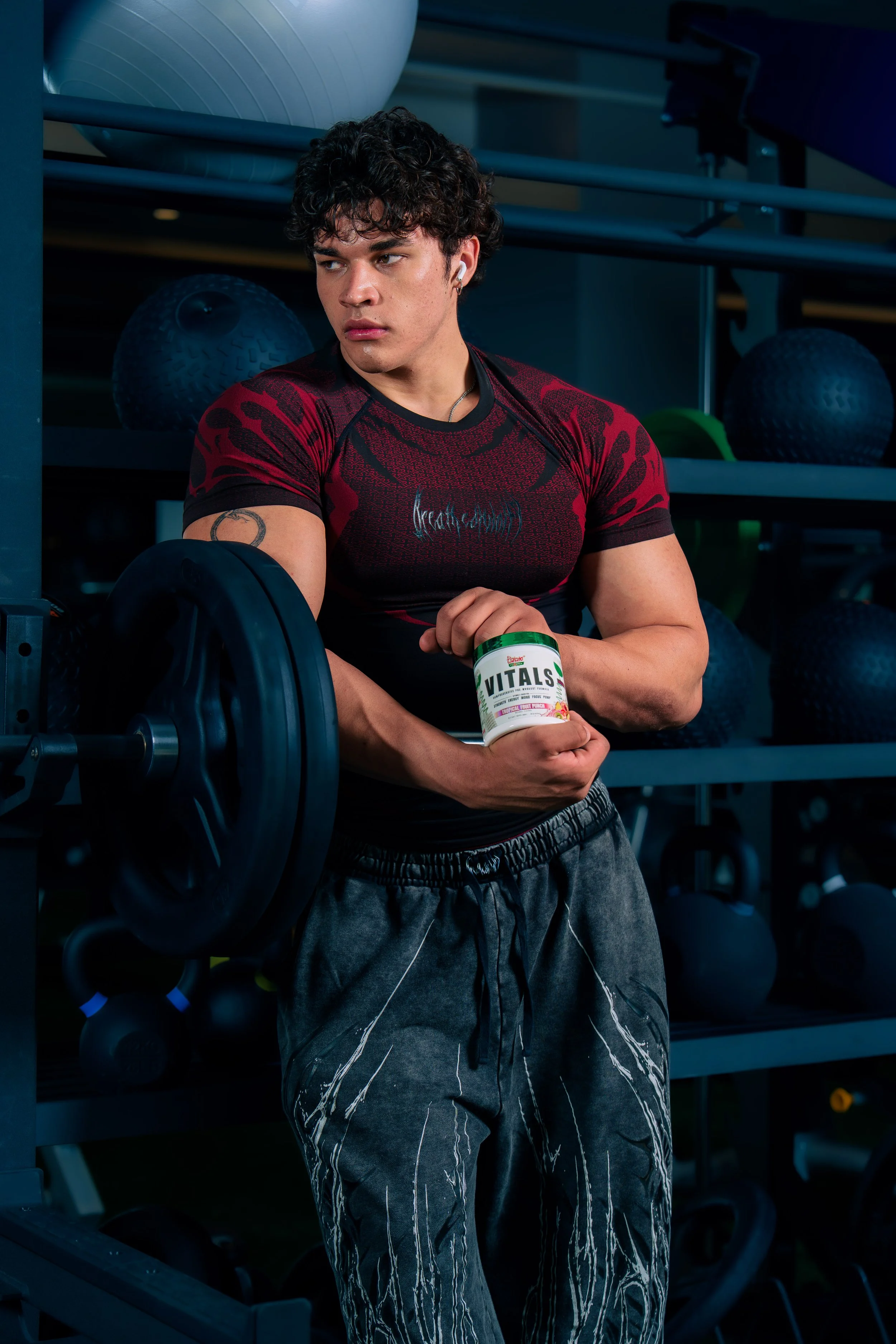A young man with curly hair, wearing a red and black athletic shirt, standing in a gym, holding a container of Vitalis dietary supplement, with gym equipment and medicine balls in the background.