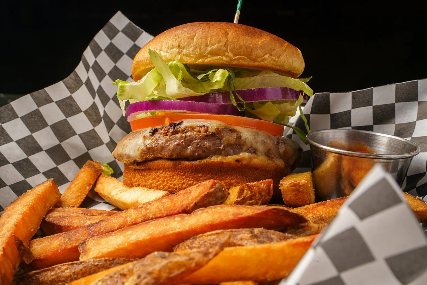 Close-up of a cheeseburger with lettuce, tomato, red onion, and pickles, served with French fries and a small container of dipping sauce on a checkered paper-lined tray.
