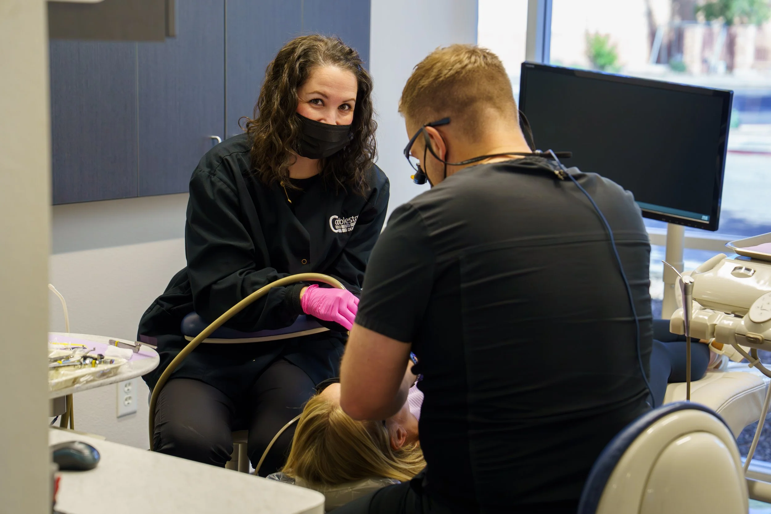 A medical professional with dark curly hair, wearing a black mask and pink gloves, is attending to a patient lying on a dental chair. Another person with short red hair and glasses is visible. The setting appears to be a dental office with a computer