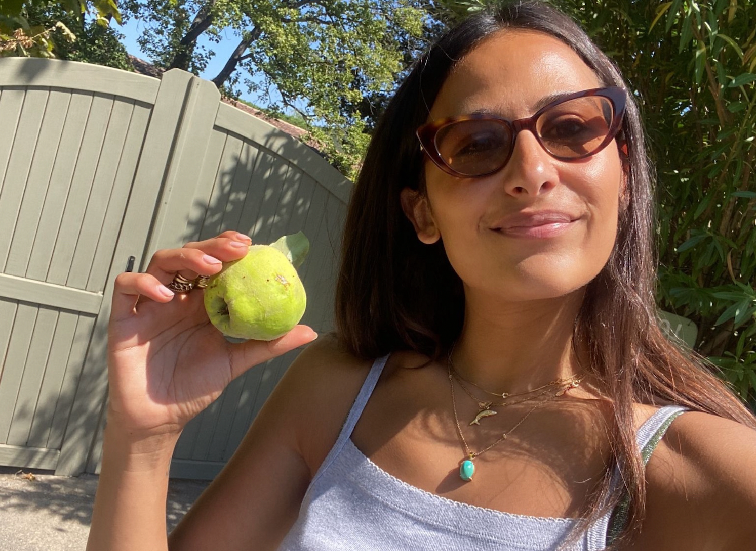 A woman with long brown hair, wearing sunglasses, a white tank top, and necklaces, holding a ripe green apple outdoors near a wooden fence and foliage, smiling at the camera in bright sunlight.