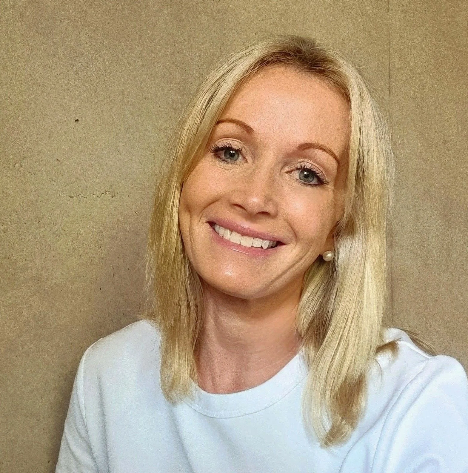 Portrait of a smiling woman with blonde hair, blue eyes, wearing pearl earrings and a white top, standing against a beige wall.