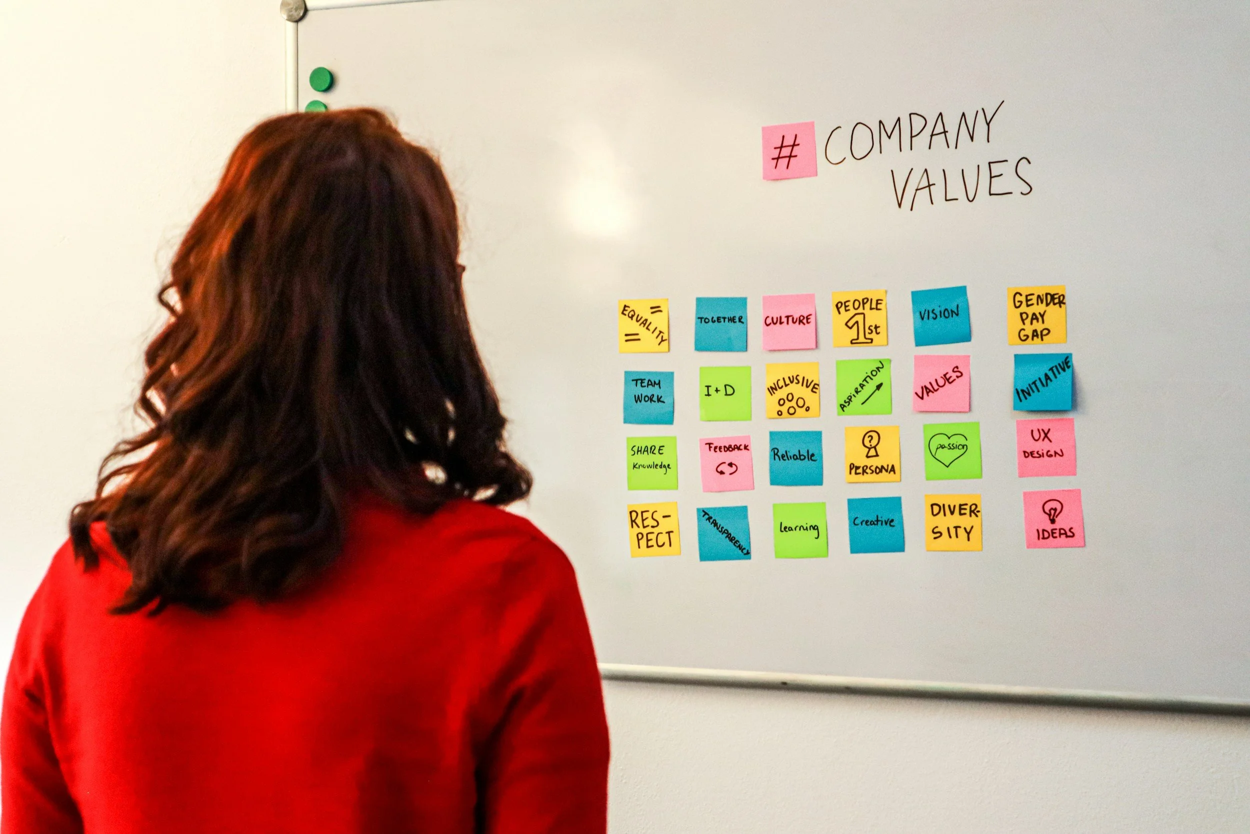 Woman with long red hair wearing a red jacket standing in front of a whiteboard with colorful sticky notes related to company values and culture.