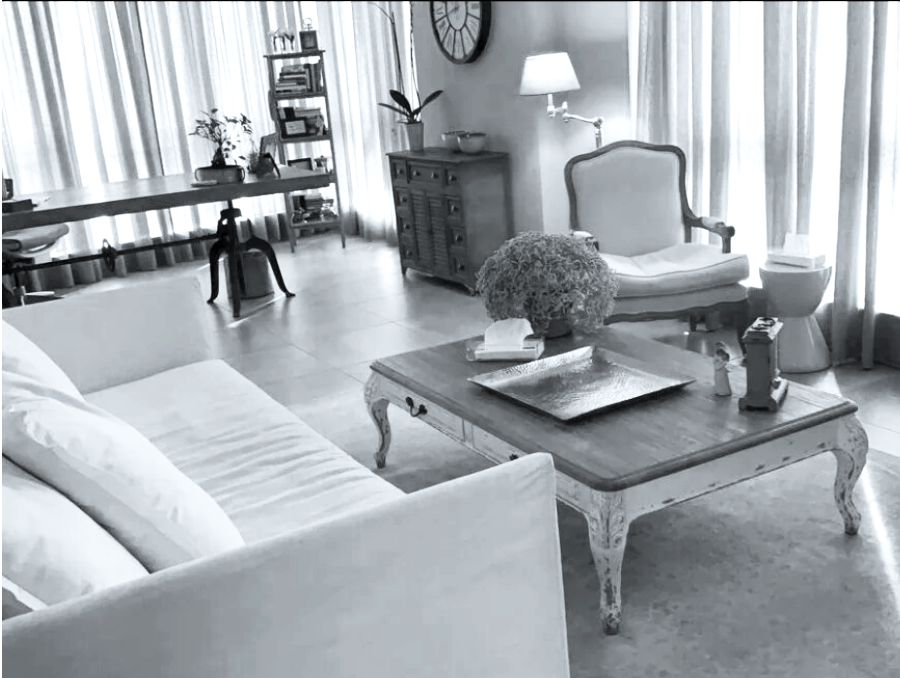 A living room with a sofa, armchair, coffee table, wall clock, lamp, bookshelf, and decorative items in a well-lit space with curtains.
