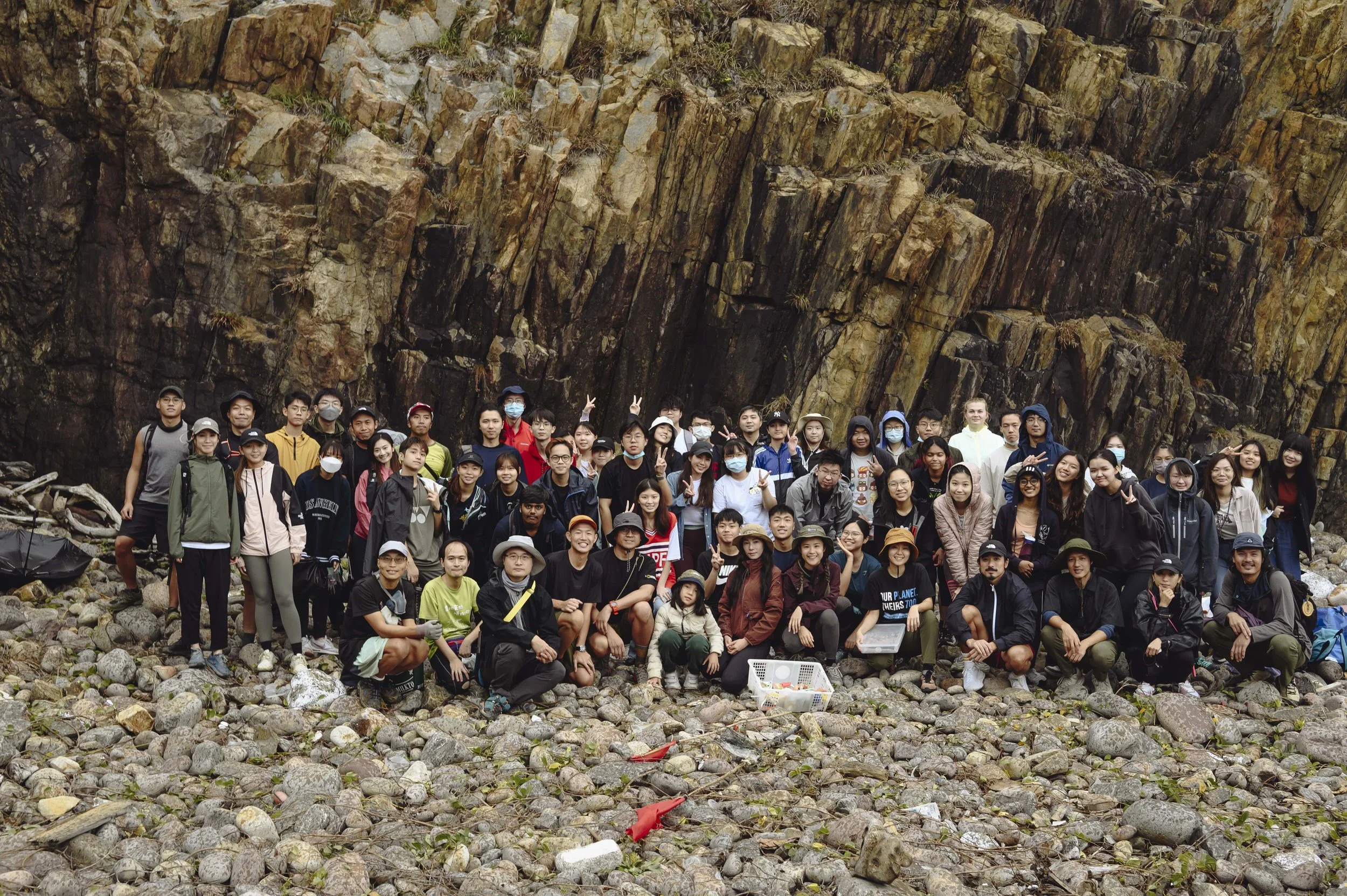 A large group of people posing for a photo on a rocky beach with large rock formations or cliffs in the background.