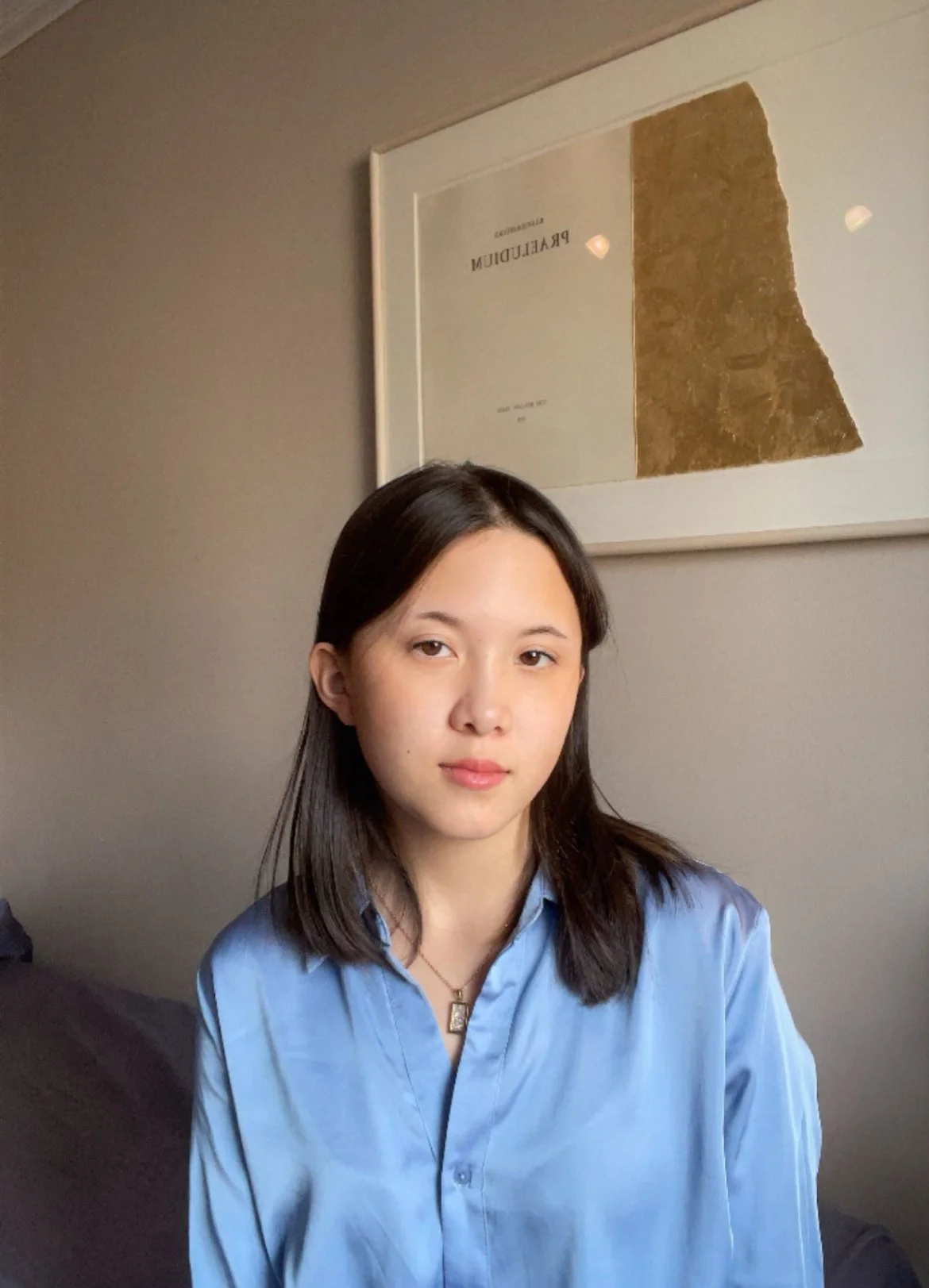 A young woman with straight dark hair, wearing a light blue button-up shirt and a necklace, sitting indoors against a gray wall with framed artwork in the background.