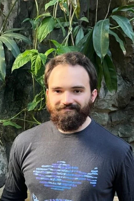 A man with dark hair, a beard, and a mustache stands in front of lush green foliage and rocks, wearing a dark gray T-shirt with a graphic design.