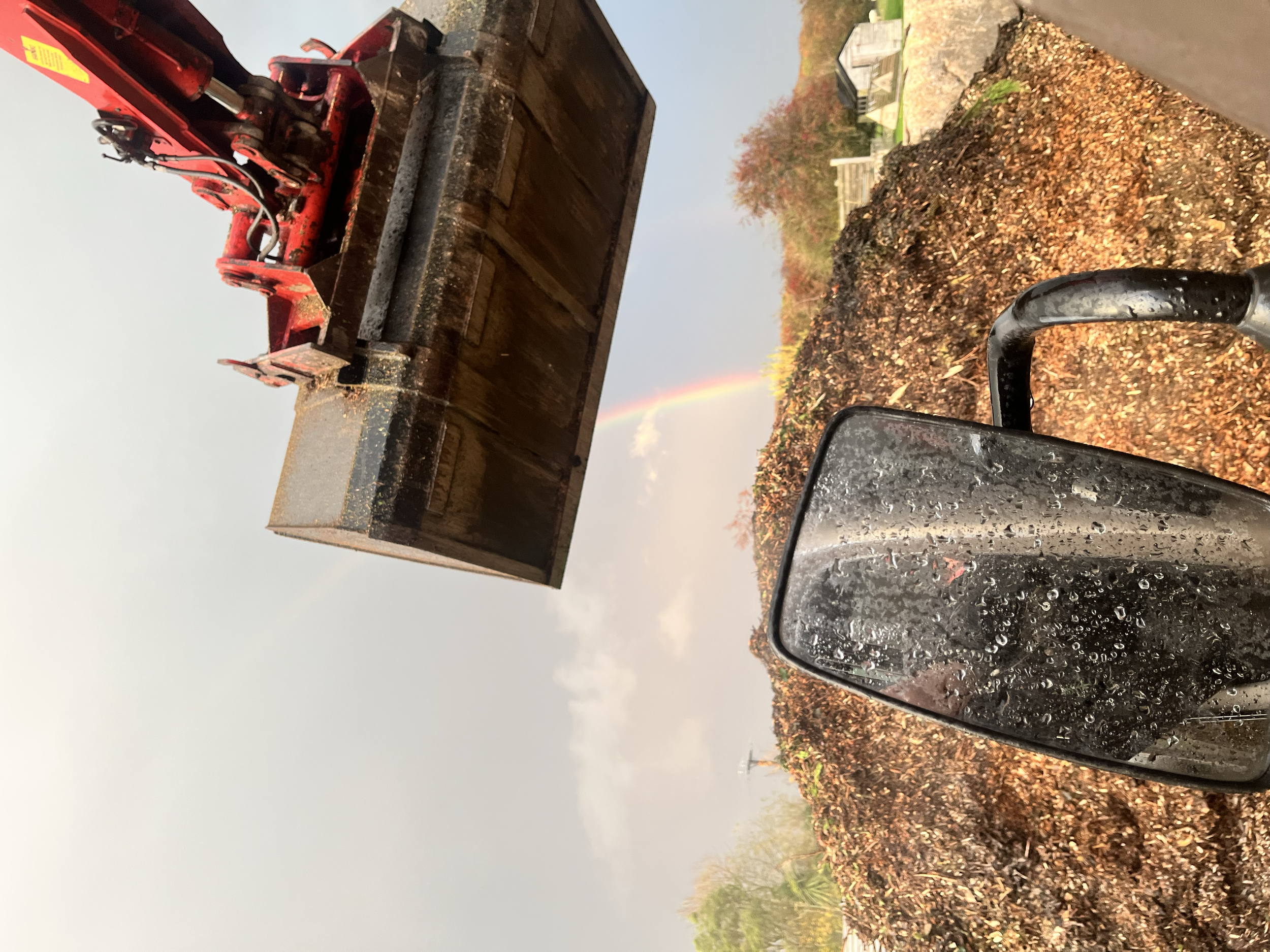 A red skid steer loader dumping mulch onto a pile. There are leaves on the ground and a rainbow in the sky.