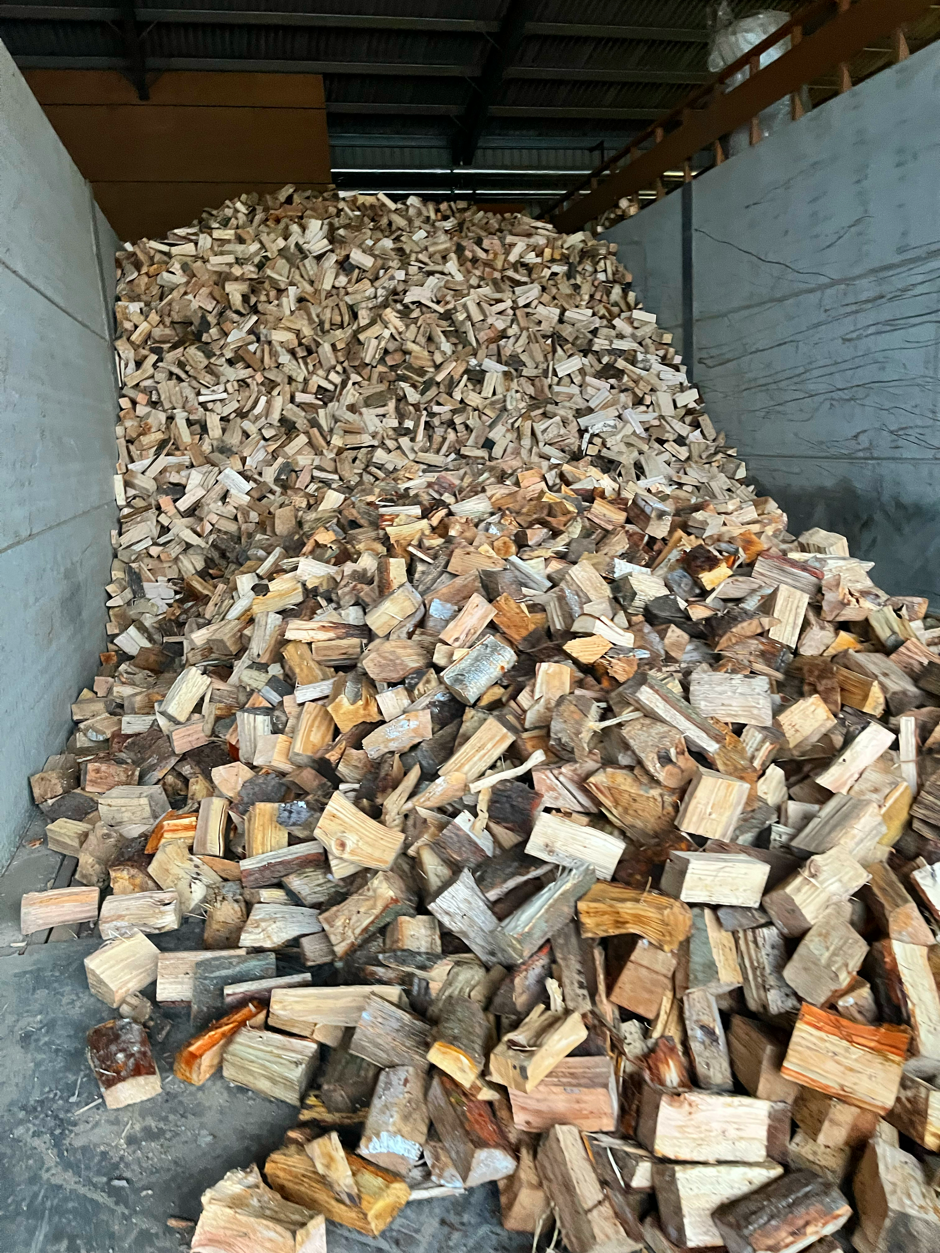 A large pile of split firewood inside a drying floor (kiln) area with a concrete wall on the left and a wooden railing on the right.