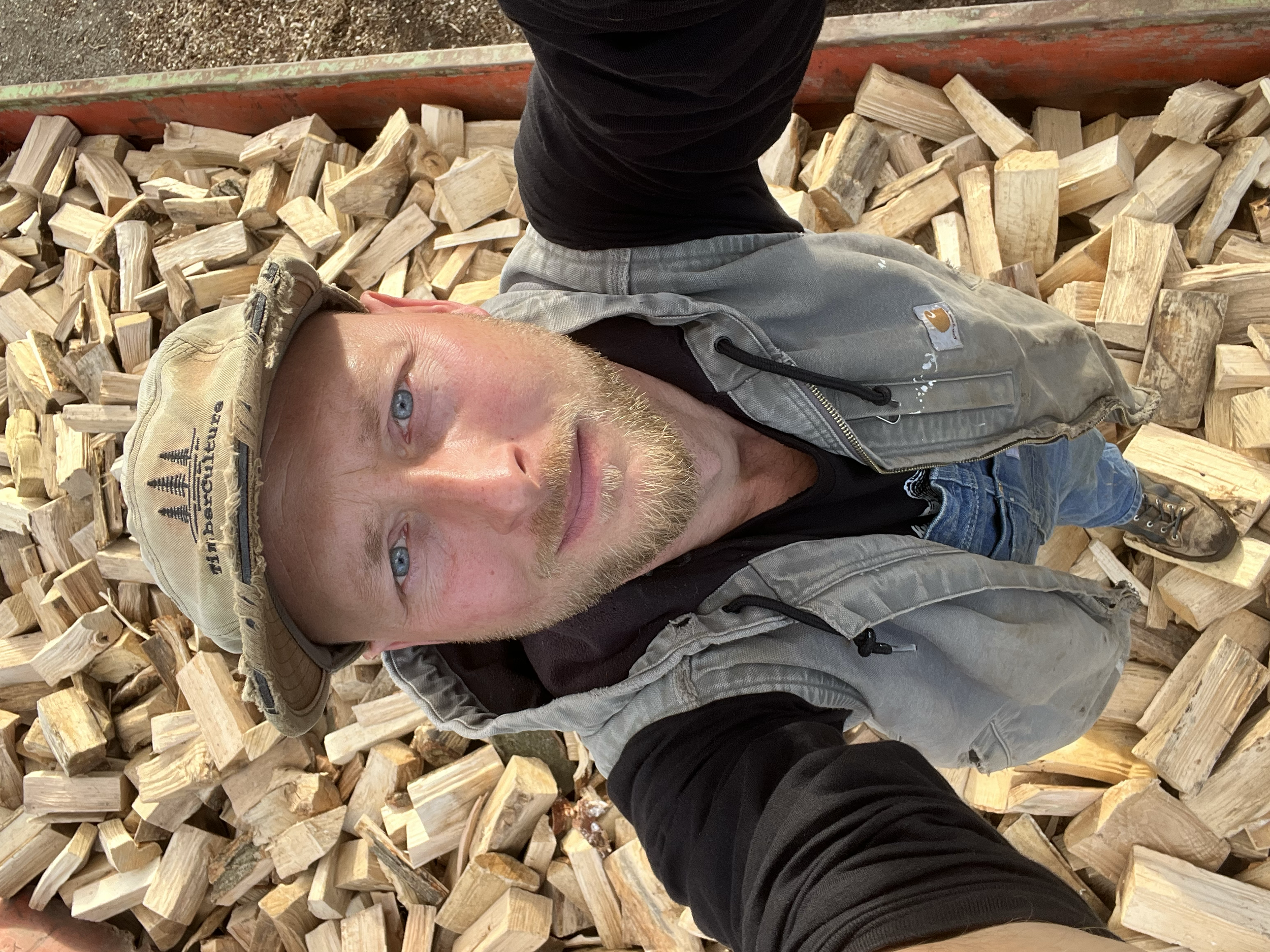A man with a blond beard and blue eyes taking a selfie from a high angle while standing on chopped firewood. He is wearing a beige Carhartt cap, a gray Carhartt vest, and a black shirt.
