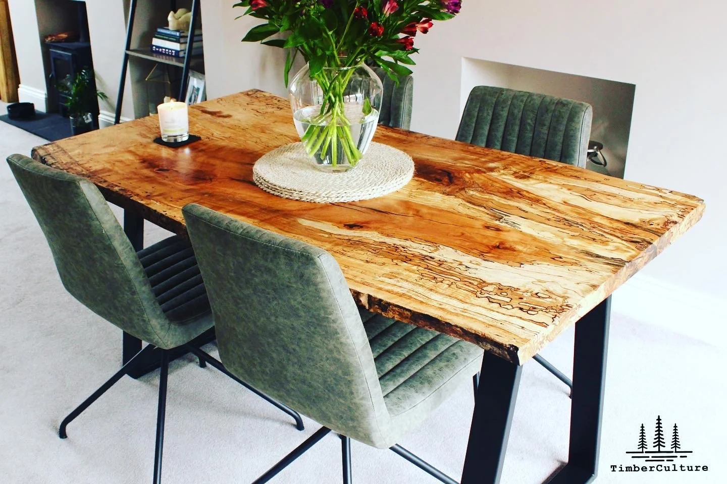 Wooden dining table with a glass vase of pink and purple flowers, a round woven placemat, and a white candle in a black holder. Four green upholstered chairs surround the table.