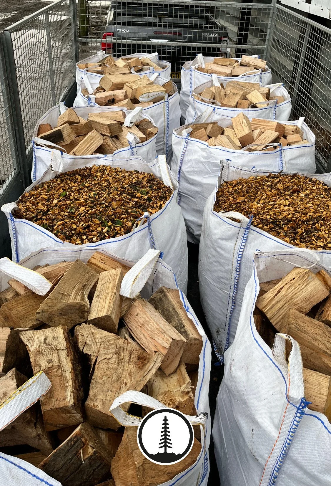Large white bags filled with chopped firewood and chopped wood logs, placed in outdoor metal fencing. A black Toyota vehicle is visible in the background.
