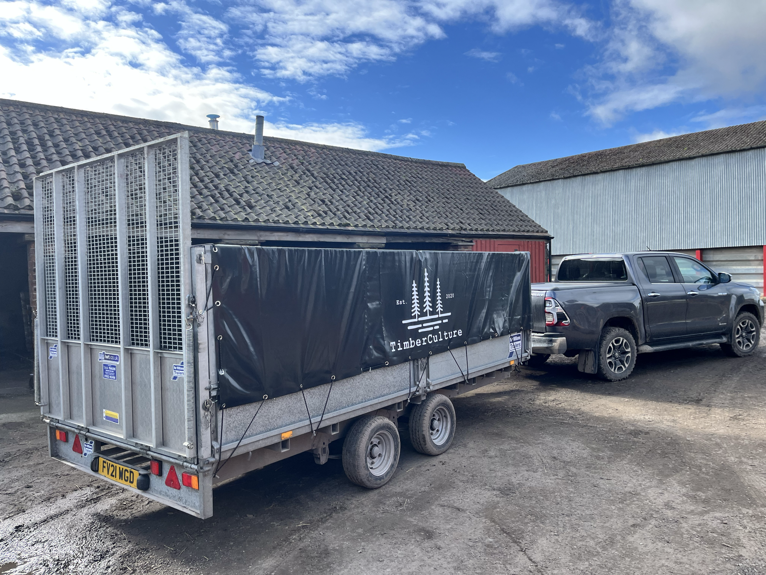 A black pickup truck parked on a dirt lot with a trailer attached, which has a black cover with white trees and the text 'TimberCulture' on it. In the background, there is a building with a tiled roof and another building with a corrugated metal exte