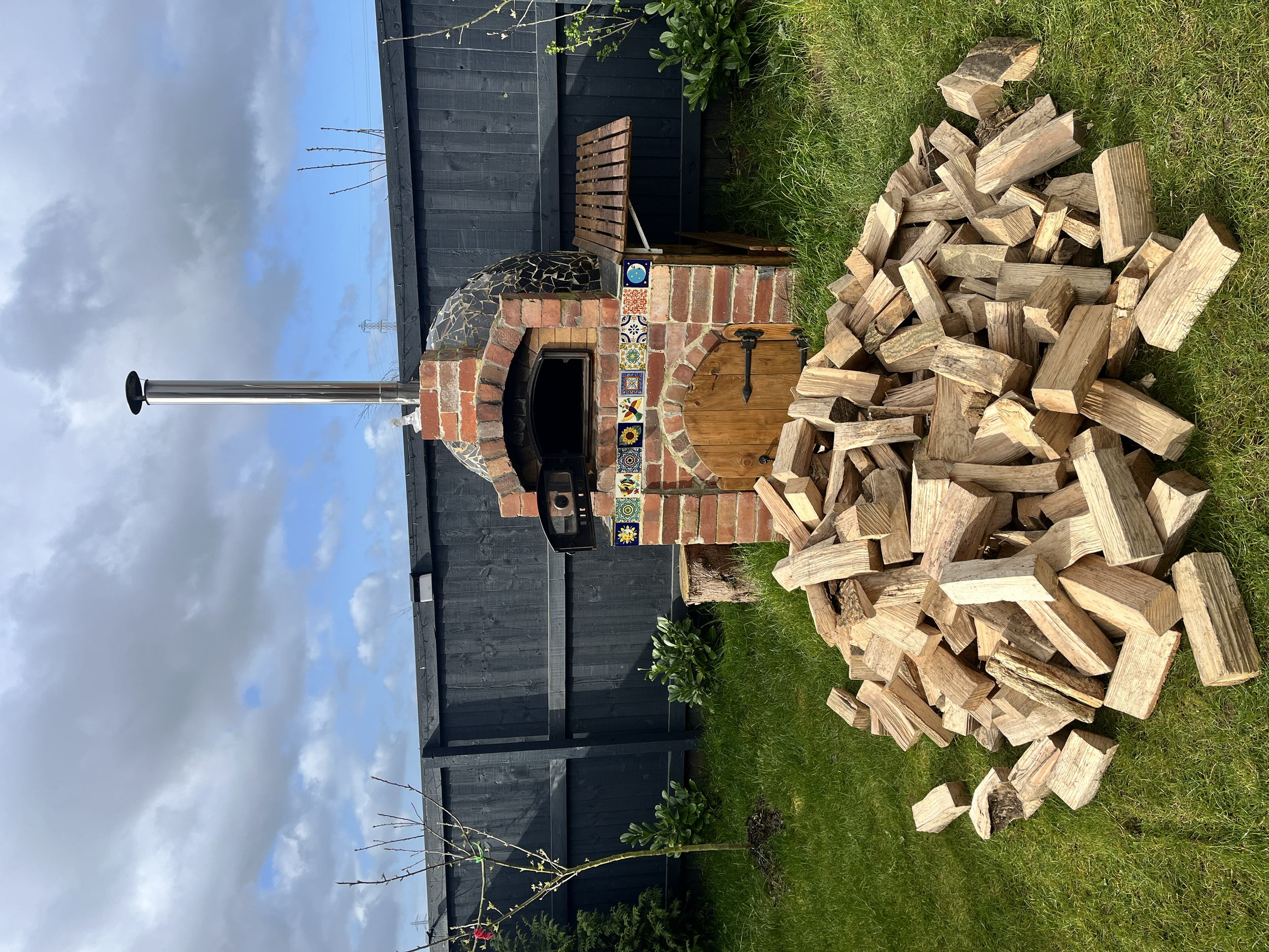 An outdoor brick pizza oven with a chimney, decorated with colorful tiles, surrounded by a black wooden fence, and a pile of chopped firewood in front.