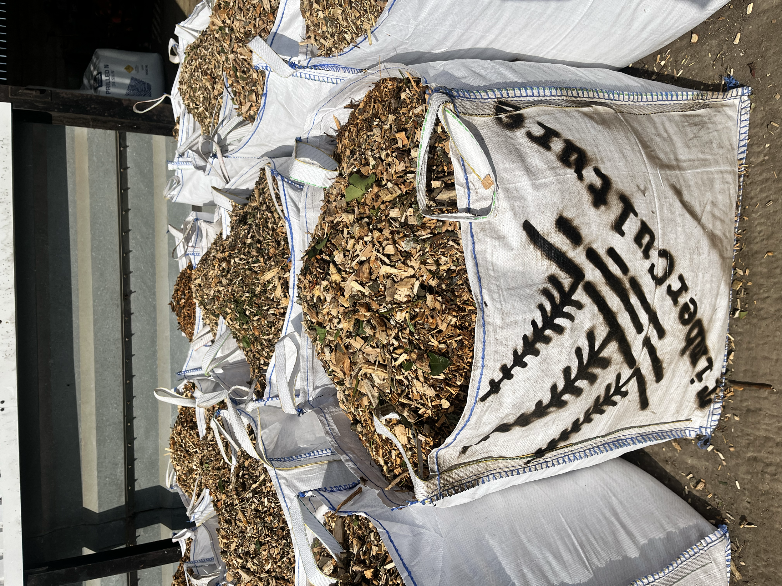 Large white bags filled with cut wood chips or bark outside, with some spilled on the ground, on a concrete surface.