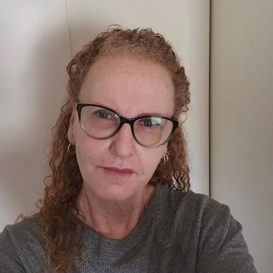 Close-up of a woman with curly red hair, wearing glasses, and small hoop earrings, looking directly at the camera.