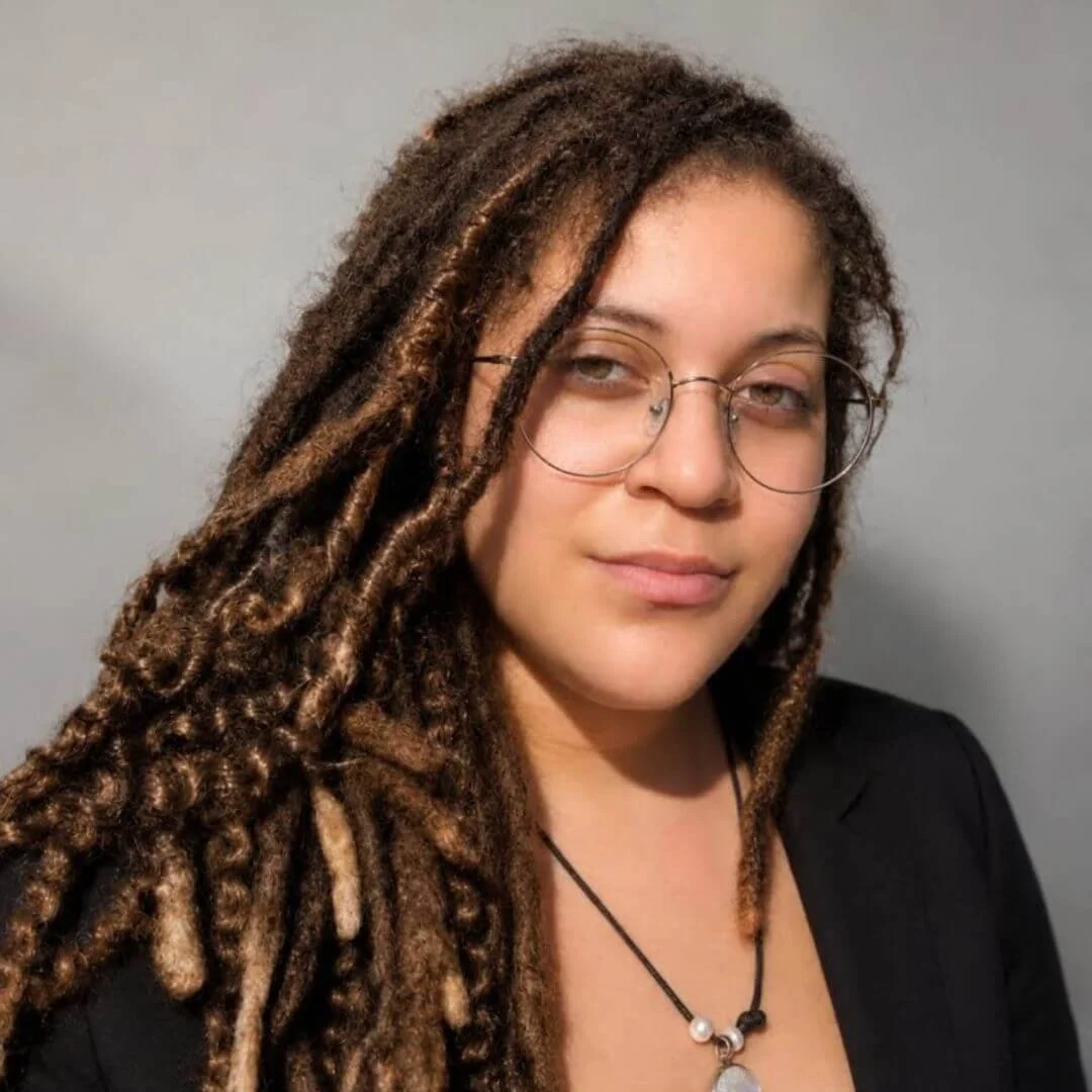 A woman with brown glasses and dreadlocks smiling with closed eyes outdoors, with a background of green trees and mountains under a cloudy sky.