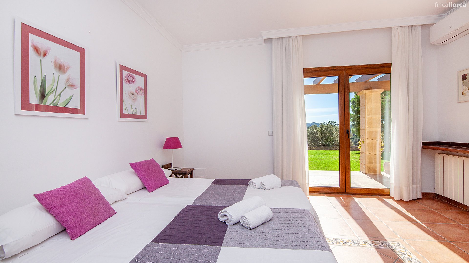 A bedroom with two beds, purple pillows, and rolled white towels, with a balcony door opening to a green outdoor area.