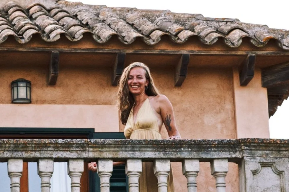 A woman with long, wavy hair wearing a yellow dress, smiling, standing on a balcony of a stucco house with a tiled roof.