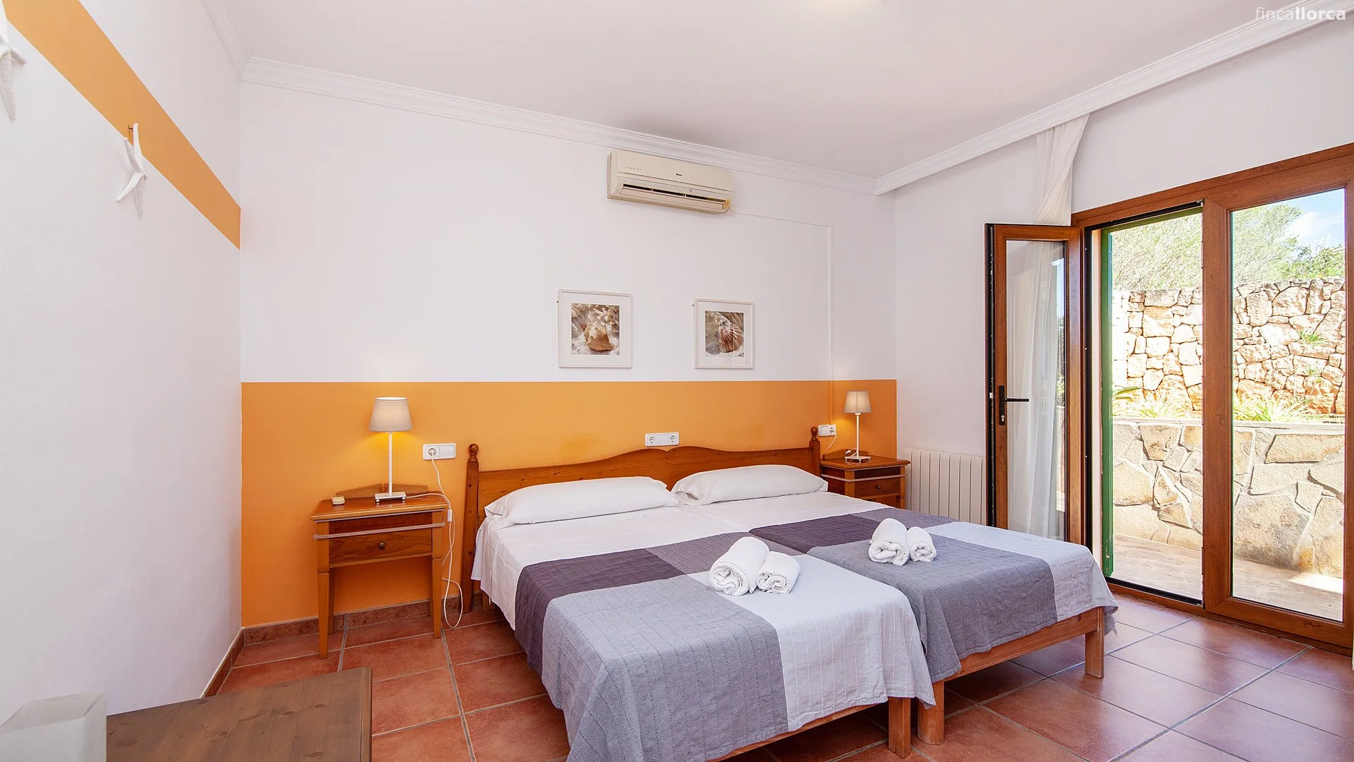 Hotel room with a double bed, two bedside tables with lamps, and an open sliding glass door leading outside to a stone patio with a stone wall in the background.