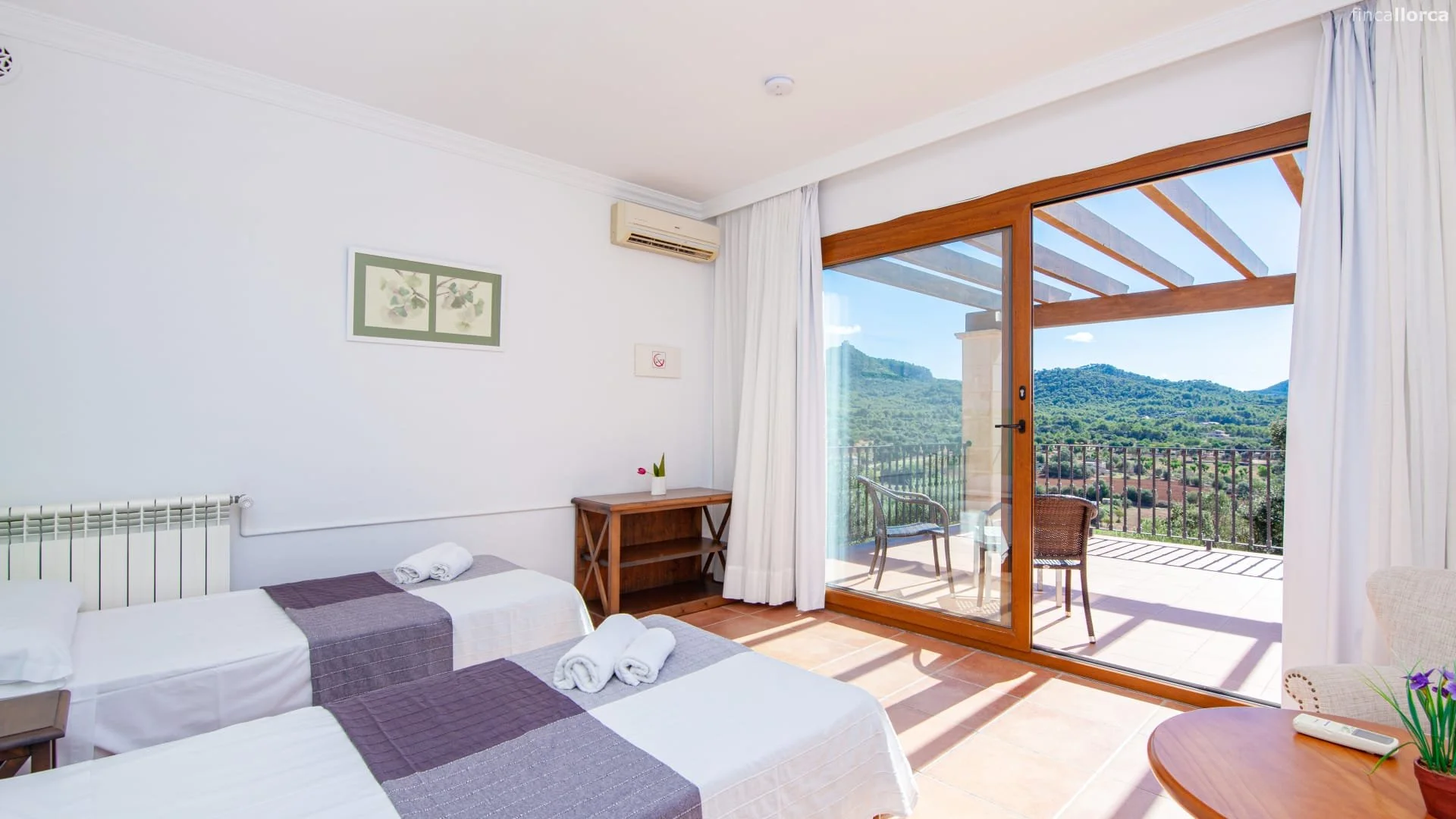 Hotel room with two single beds, overlooking a balcony with patio chairs and a scenic view of mountains and greenery.