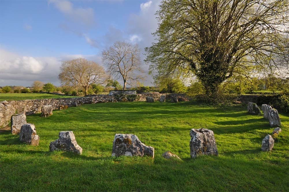 Stone circle on a grassy field with large trees and a stone wall in the background under a partly cloudy sky.