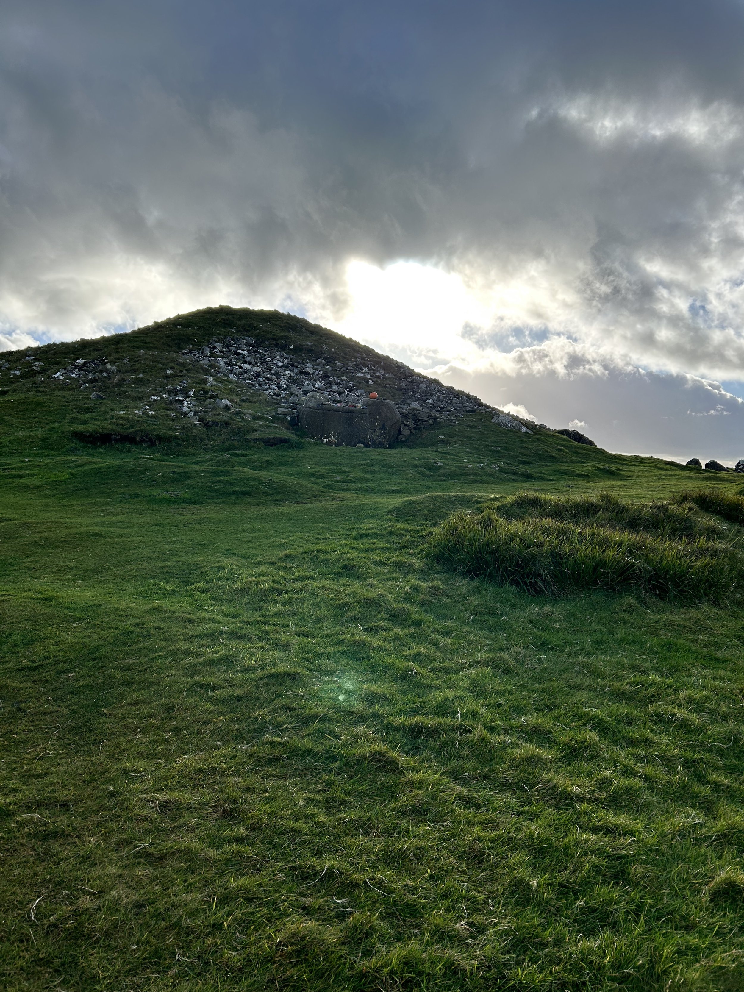 Grassy hill with rocky mound and cloudy sky, sunlight breaking through.