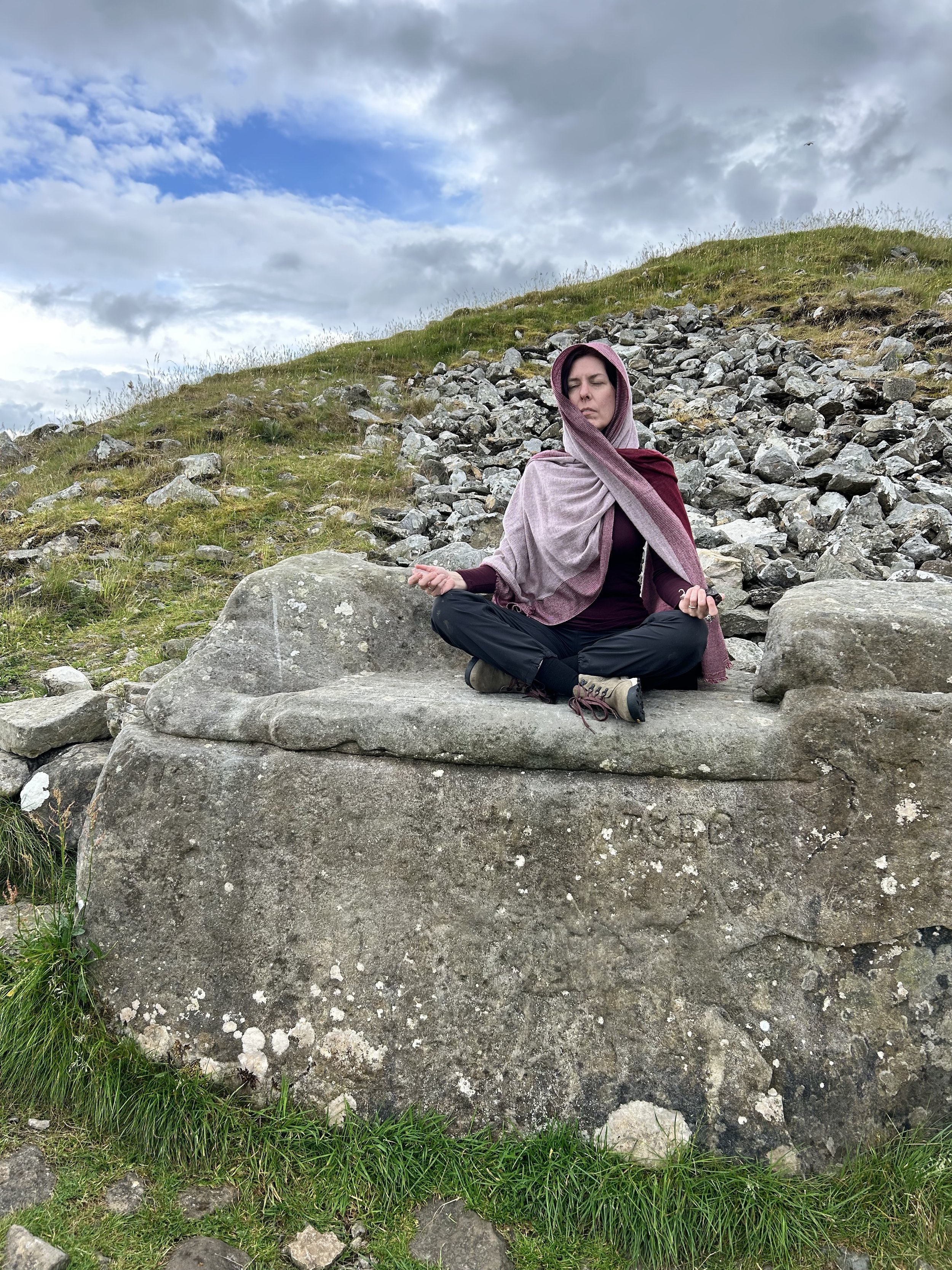 Person meditating on a large stone in a rocky landscape under a cloudy sky.